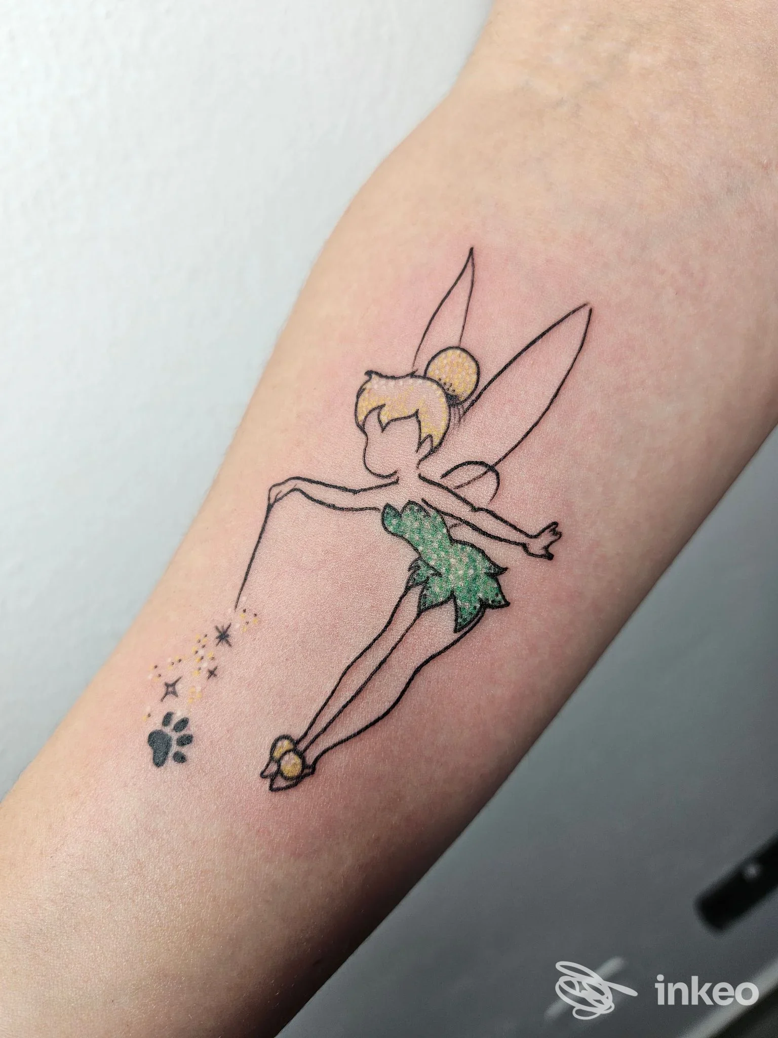 Tatuaggio su avambraccio: una fata in verde con ali, in volo, con bacchetta che sprigiona piccole stelle.