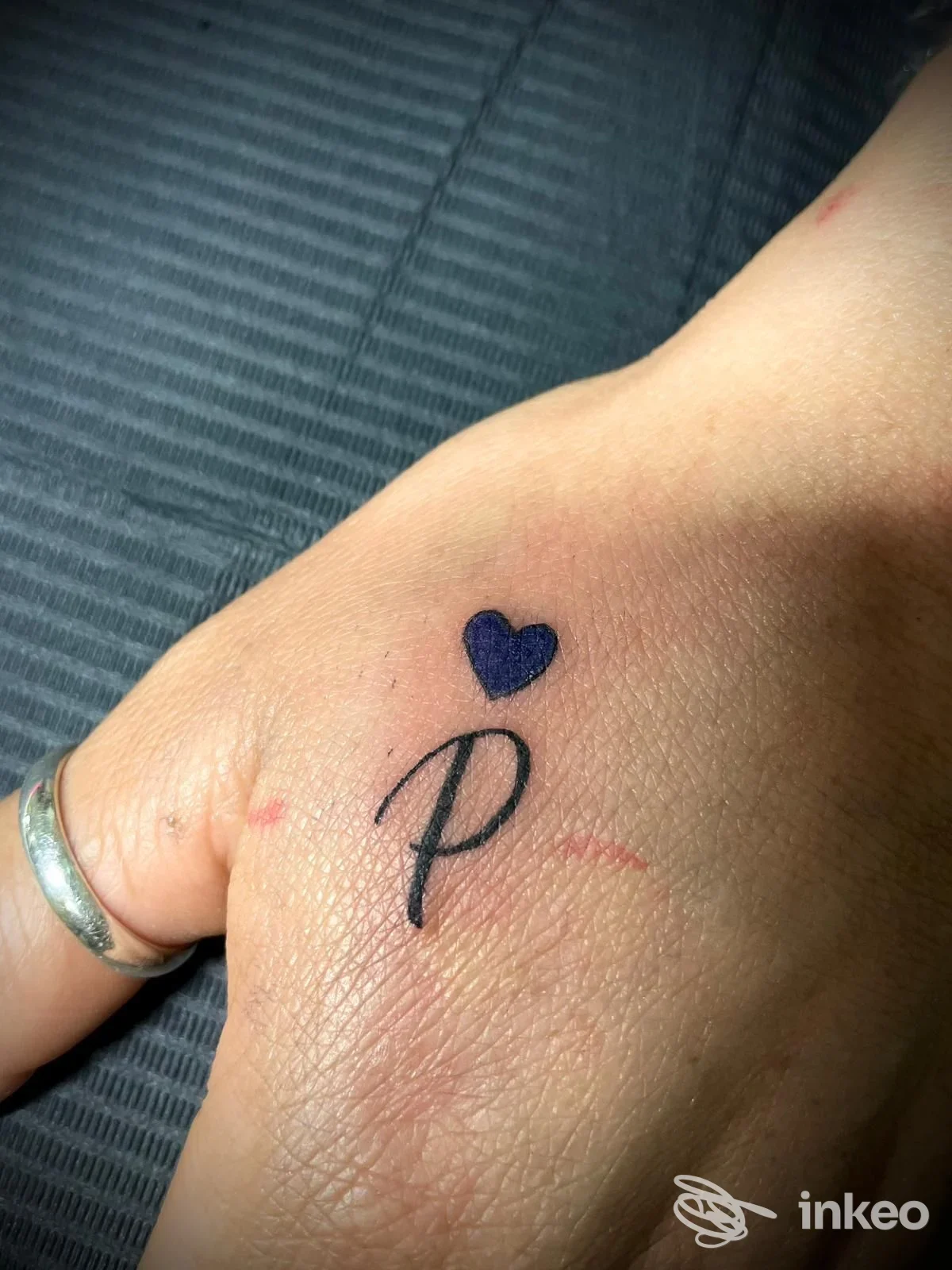 Piccolo tatuaggio sul dorso della mano con una P in corsivo e un cuore viola, foto per Clara Longo Studio - Empire Tattoo 1995, Maglie.