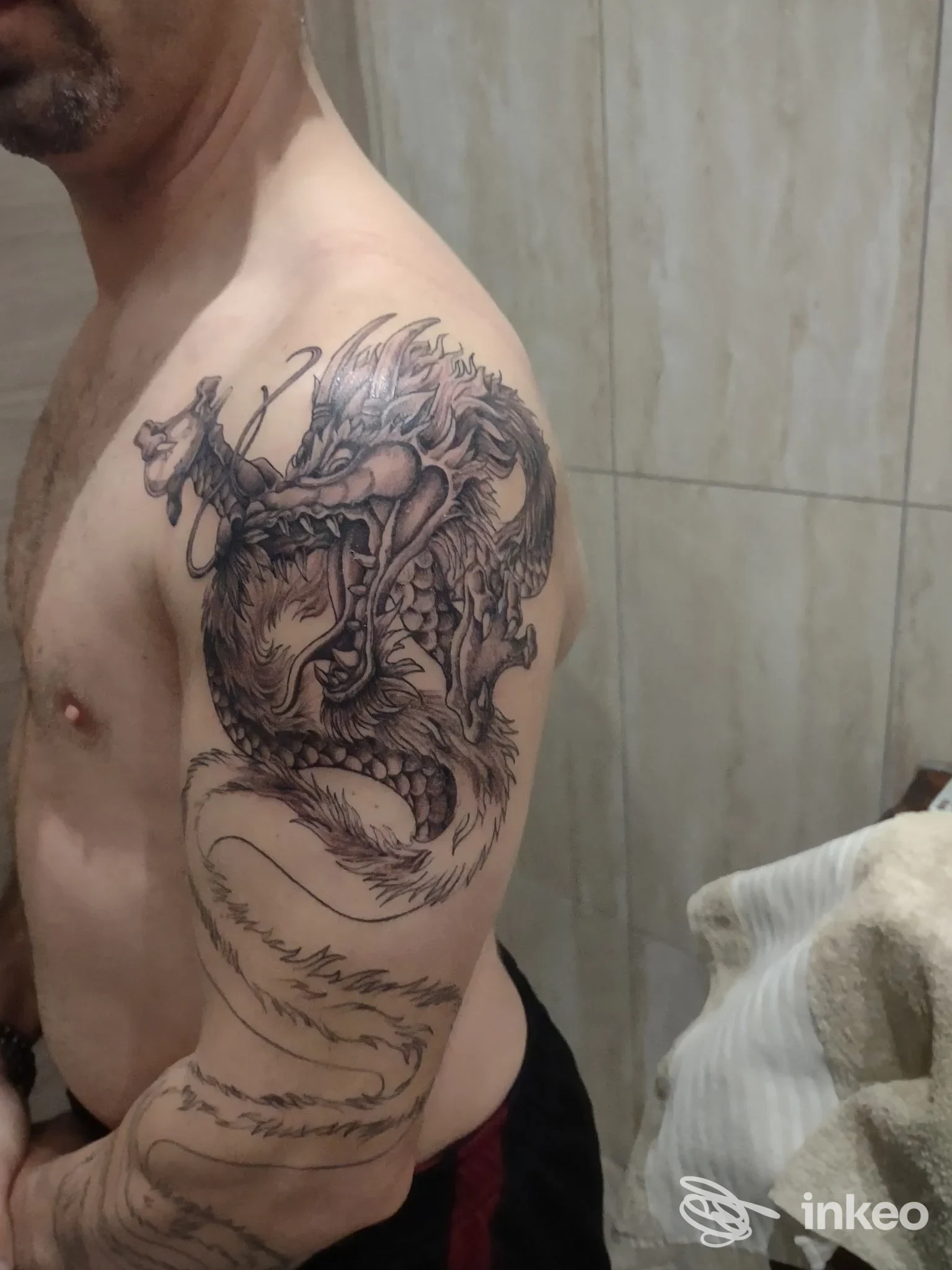 Dragon tattoo sulla spalla e sul braccio sinistro di un uomo, tatuaggio visibile su pelle nuda in ambiente di studio.