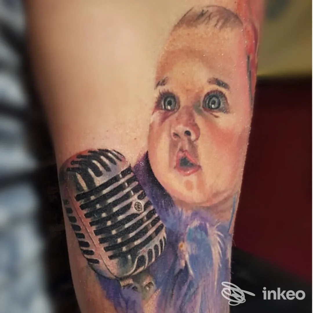Tatuaggio realistico su pelle raffigurante una neonata con espressione stupita di fronte a un microfono vintage, eseguito sul braccio. Studio Pride & Glory Tattoo.