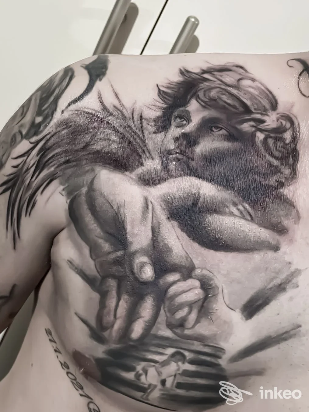 Grande tatuaggio realistico in bianco e nero su petto e parte superiore del torace, angelo con ali e mano; immagine per Ink Warriors Tattoo Studio, Manduria.