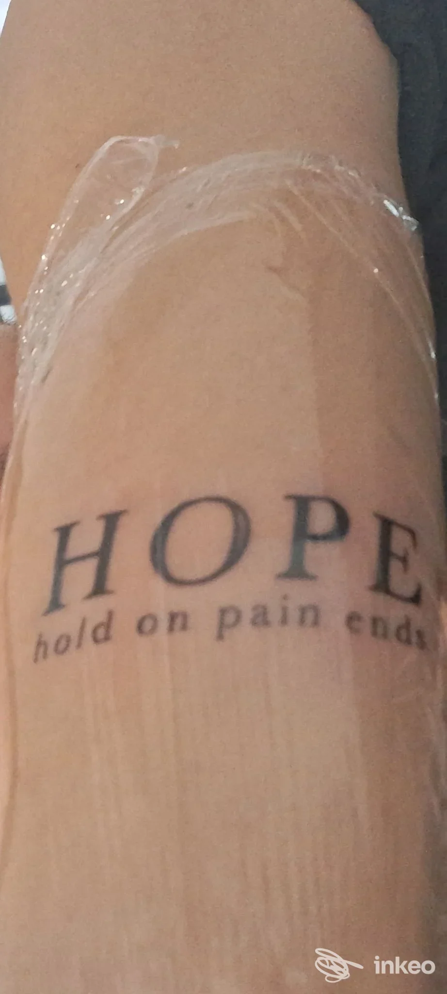 Braccio con tatuaggio visibile che legge HOPE e la frase sottostante, con la pelle avvolta da pellicola trasparente; nessuna persona in primo piano.