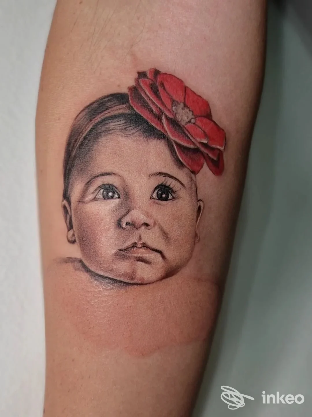 Tatuaggio realistico di un volto di neonato con fiore rosso sull'avambraccio interno, realizzato da INKLAM TATTOO STUDIO di Lamanna Fabio, Canosa di Puglia.