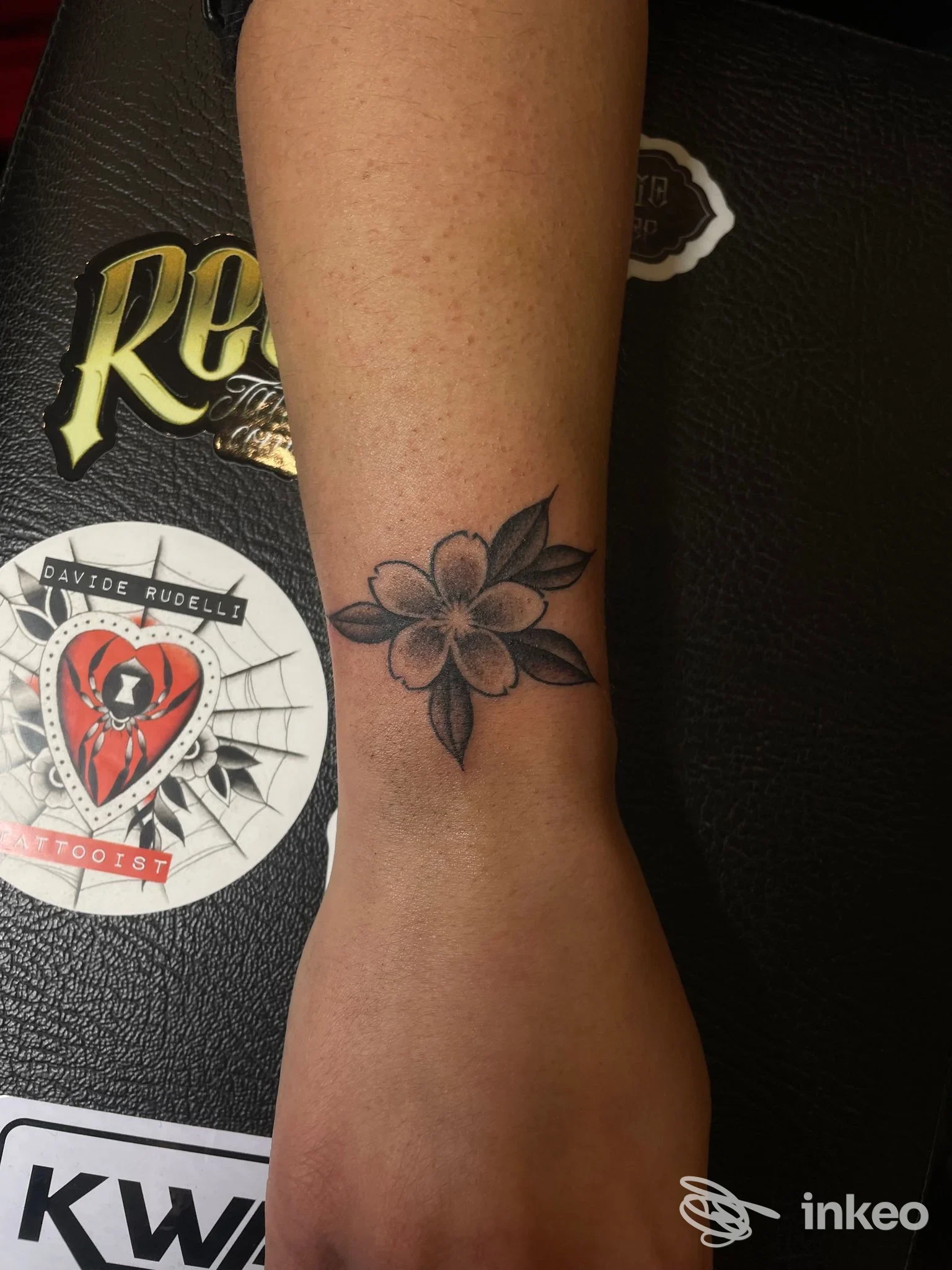 Tatuaggio floreale visibile sulla pelle del polso/avambraccio, linee nette e sfumature leggere in tonalità scure.
