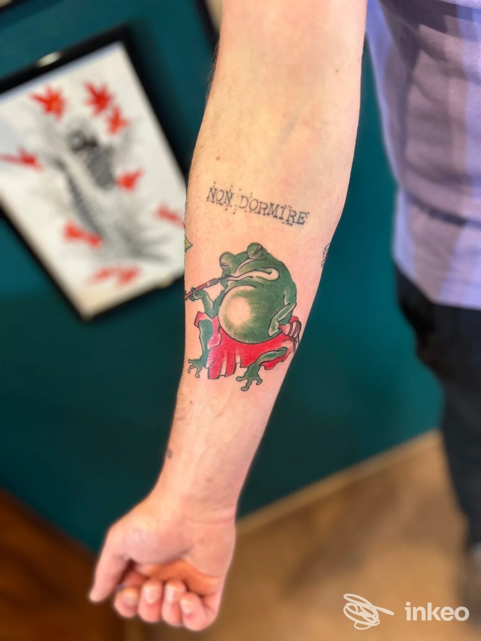 Avambraccio visibile con tatuaggio: una rana verde con zampe rosse, sopra il disegno fa capolino una dicitura scritta. Il contorno è nitido e i colori ben definiti.