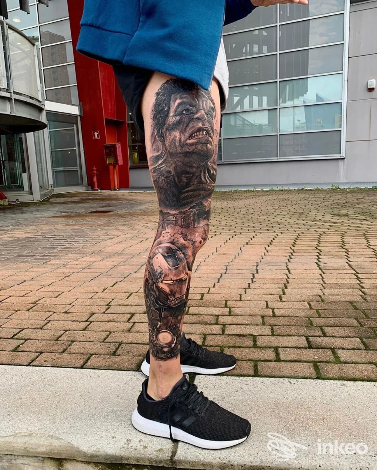 Gamba sinistra con ampio tatuaggio realistico in bianco e nero, visibile all’esterno, senza volti interi nella scena, scatto singolo davanti a edificio moderno, nessuna interfaccia sociale.