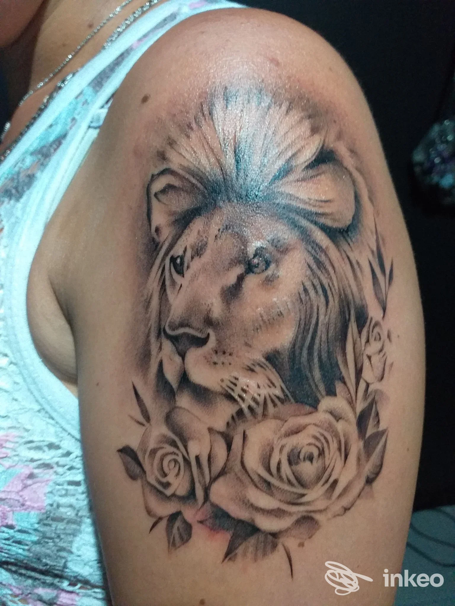 Tattoo realistico su braccio: leone stilizzato circondato da rose e foglie, realizzato in tonalità grigio-nero, con dettagli sfumati e lucidi.