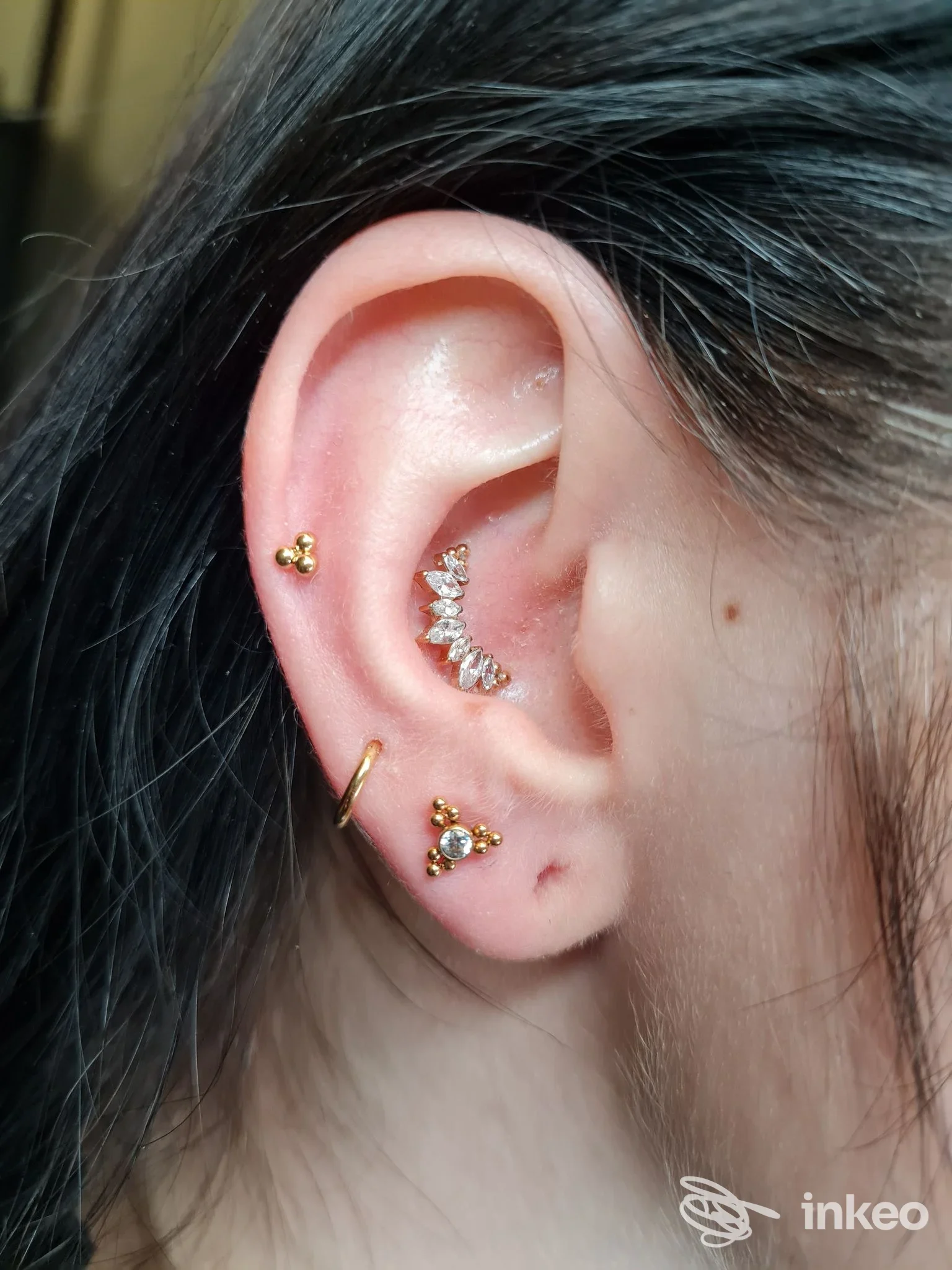 Orecchio sinistro con piercing multipli: due gioielli dorati al lobo e una fila di zirconi lungo la cartilagine interna, scatto ravvicinato.