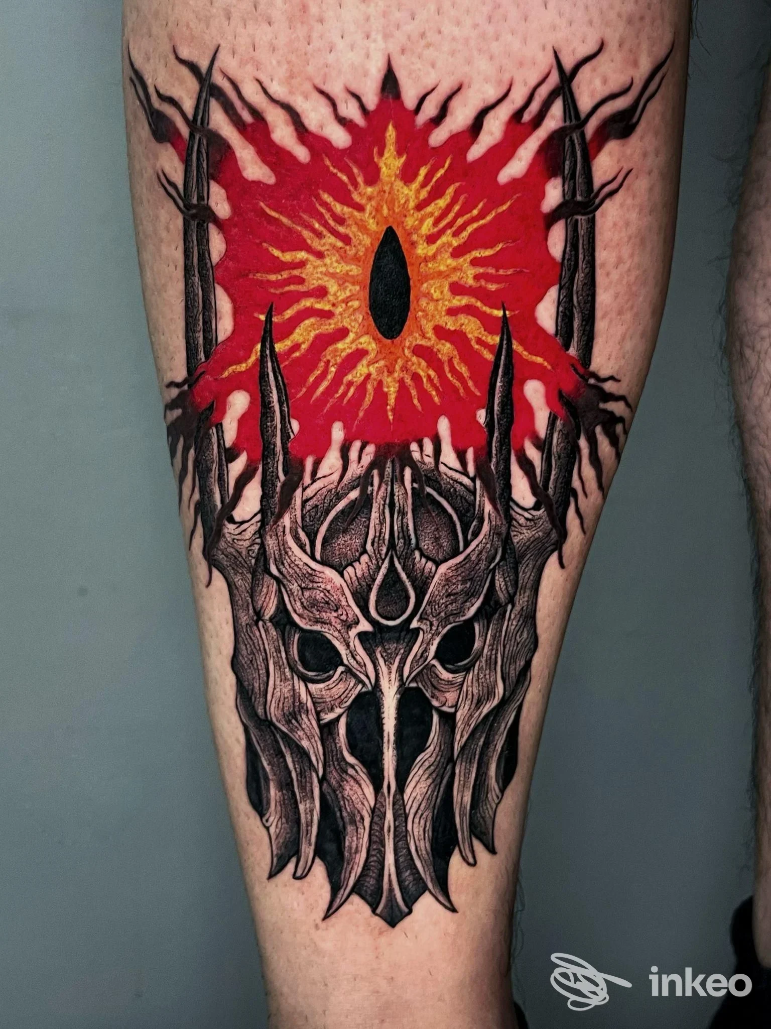 Tatuaggio su polpaccio con maschera stilizzata nera e simbolo solare rosso/giallo, foto fornita da Grass Bad Tattoo, Bari.