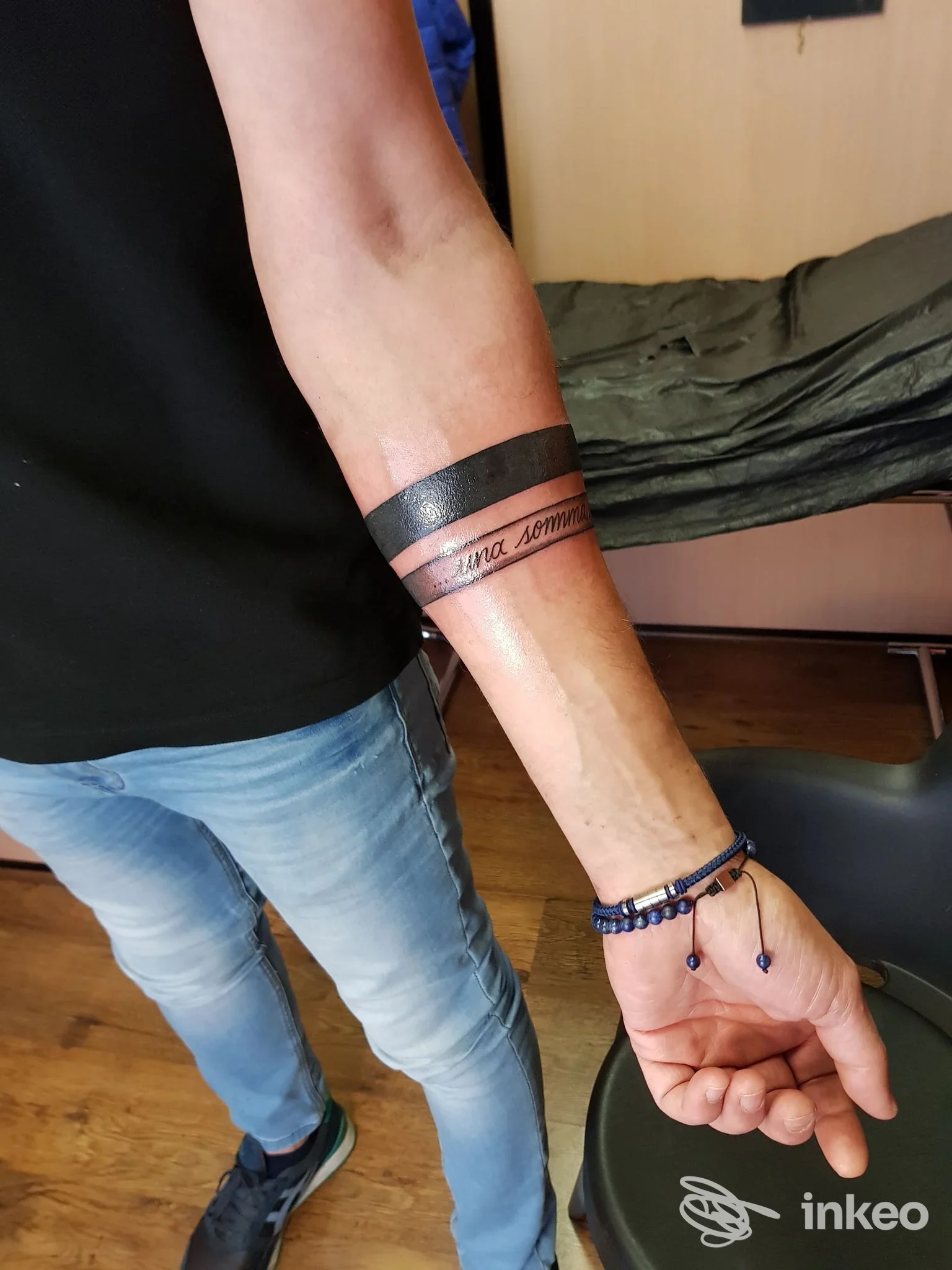 Braccio di una persona con un tatuaggio a banda nera larga attorno al polso, con una scritta sottile all'interno della banda. sfondo di letto e pavimento in legno.