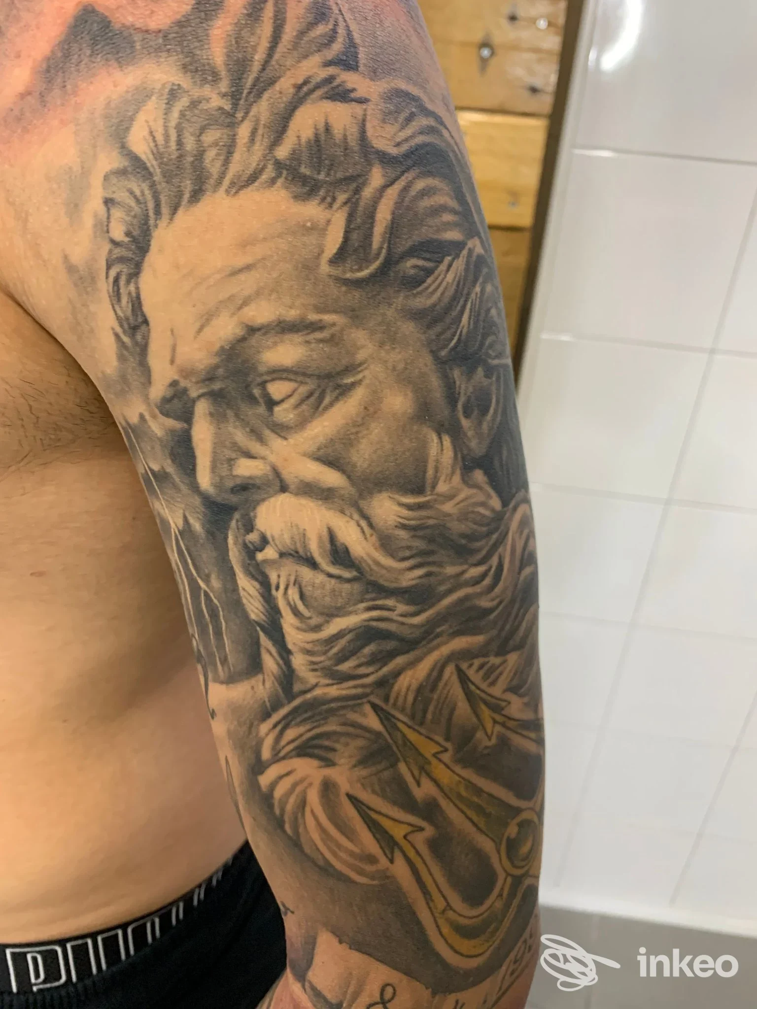 Mezza manica realistica in bianco e nero con ritratto maschile e tridente dorato sul braccio; immagine per Old Fishermen Tattoo, Martina Franca.