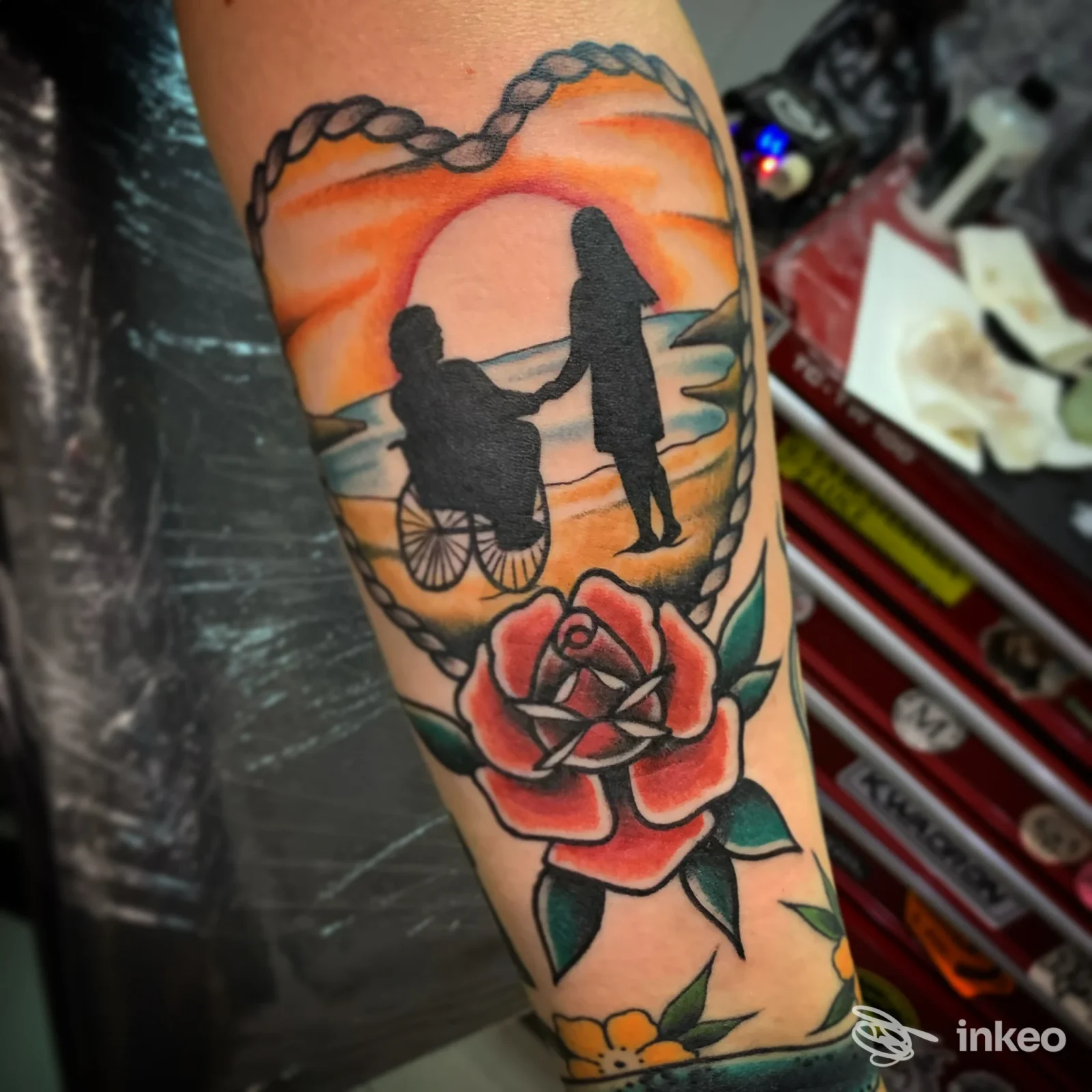 Tatuaggio su avambraccio con silhouette di persona in carrozzina e accompagnatrice davanti al tramonto e una rosa, Old Fishermen Tattoo, Martina Franca.
