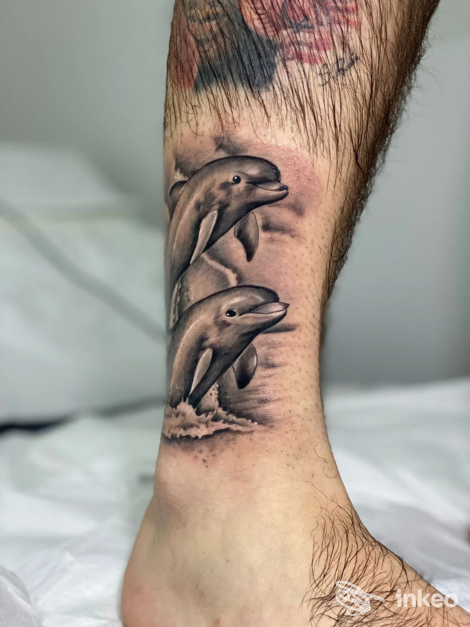 Tatuaggio realistico di due delfini sulla caviglia, lavoro di Steven Tattoo Artist a Mesagne, sfumature in bianco e nero con dettagli d'acqua.