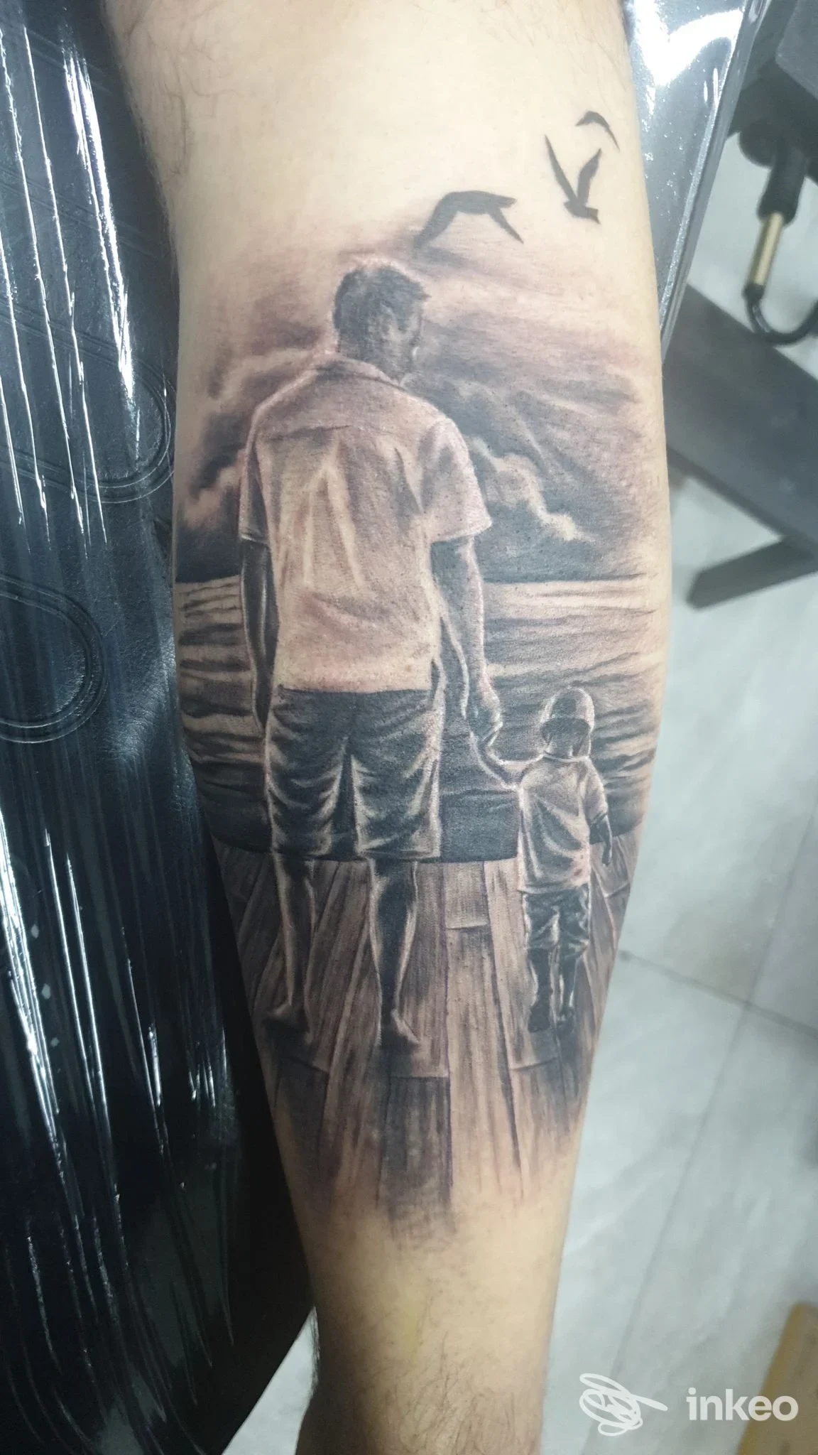 Tatuaggio realistico in bianco e nero su avambraccio raffigurante un uomo e un bambino su una banchina, Tattoo Pusher, Barletta.