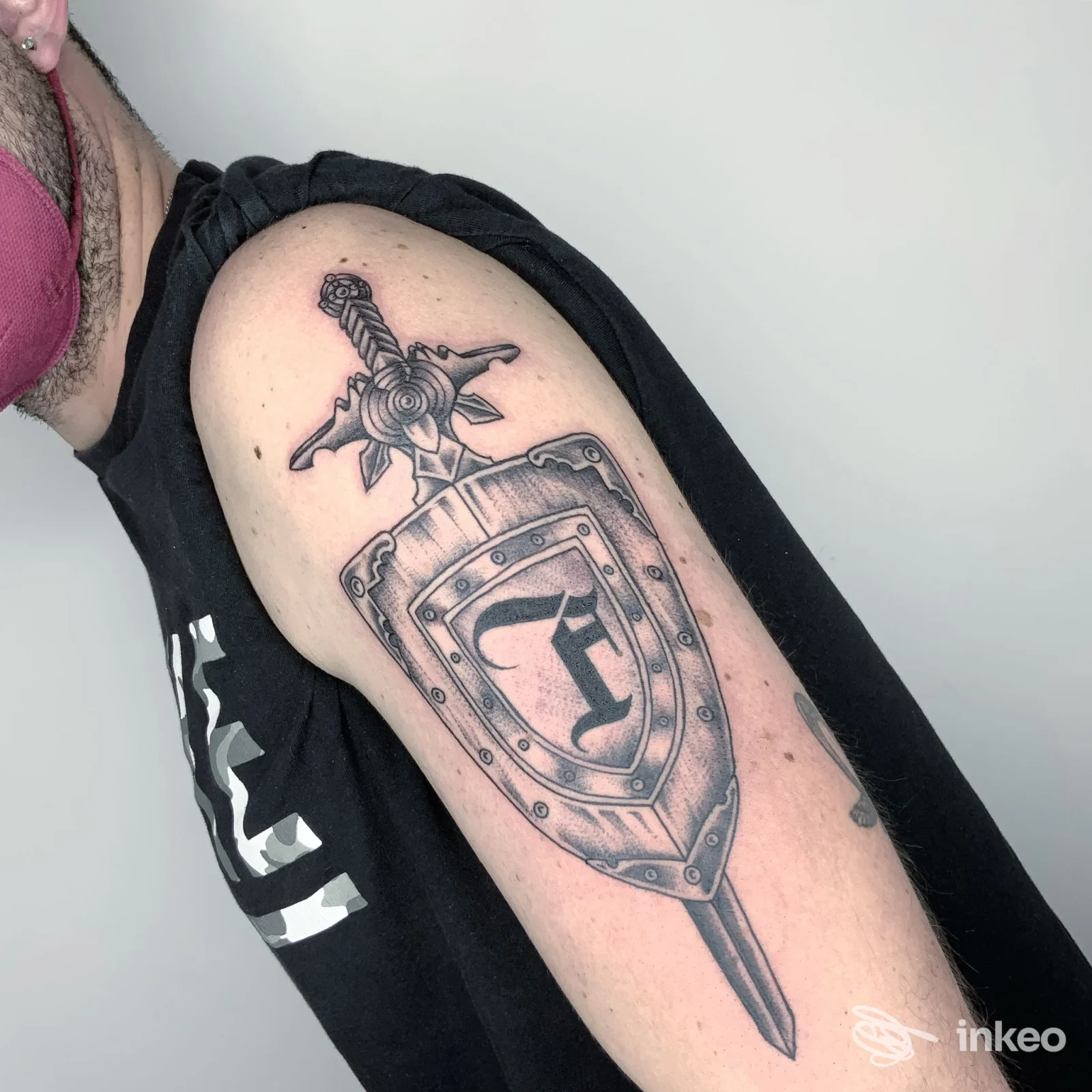 Tatuaggio a forma di scudo con spada sul deltoide, foto fornita da Tarantula Tattoo Studio, Accadia.