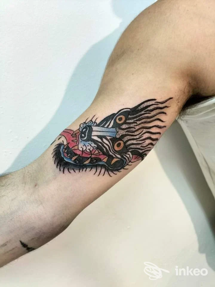 Tatuaggio colorato su interno bicipite fotografato per Battle Tattoo, Cagliari: testa stilizzata con pugnale e dettagli neri e rossi.