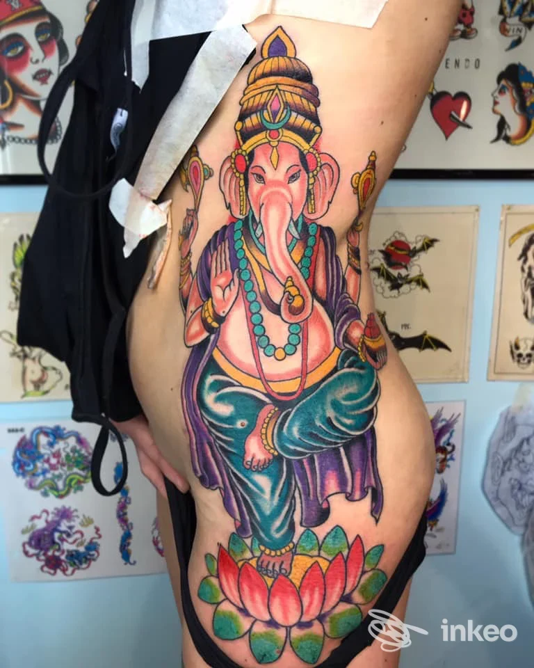 Tattoo colorato di Ganesh su coscia destra di una persona, con sfondo di pareti decorate da schizzi di tatuaggi.