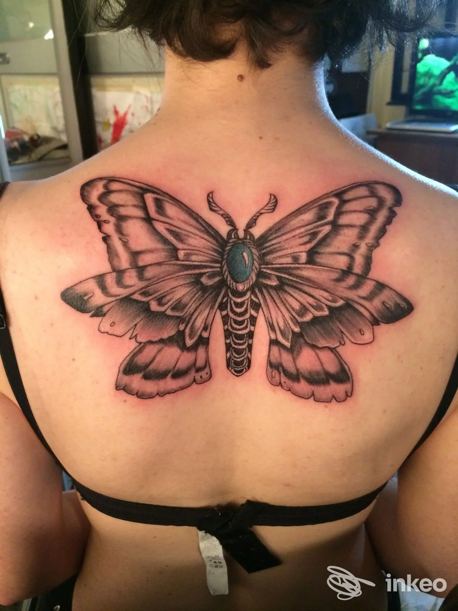 Tattoo raffigurante una farfalla nero-grigio sulla parte alta della schiena, con ali ampie e una pietra azzurra al centro; la pelle circostante è visibile e non si vedono volti o elementi promozionali.