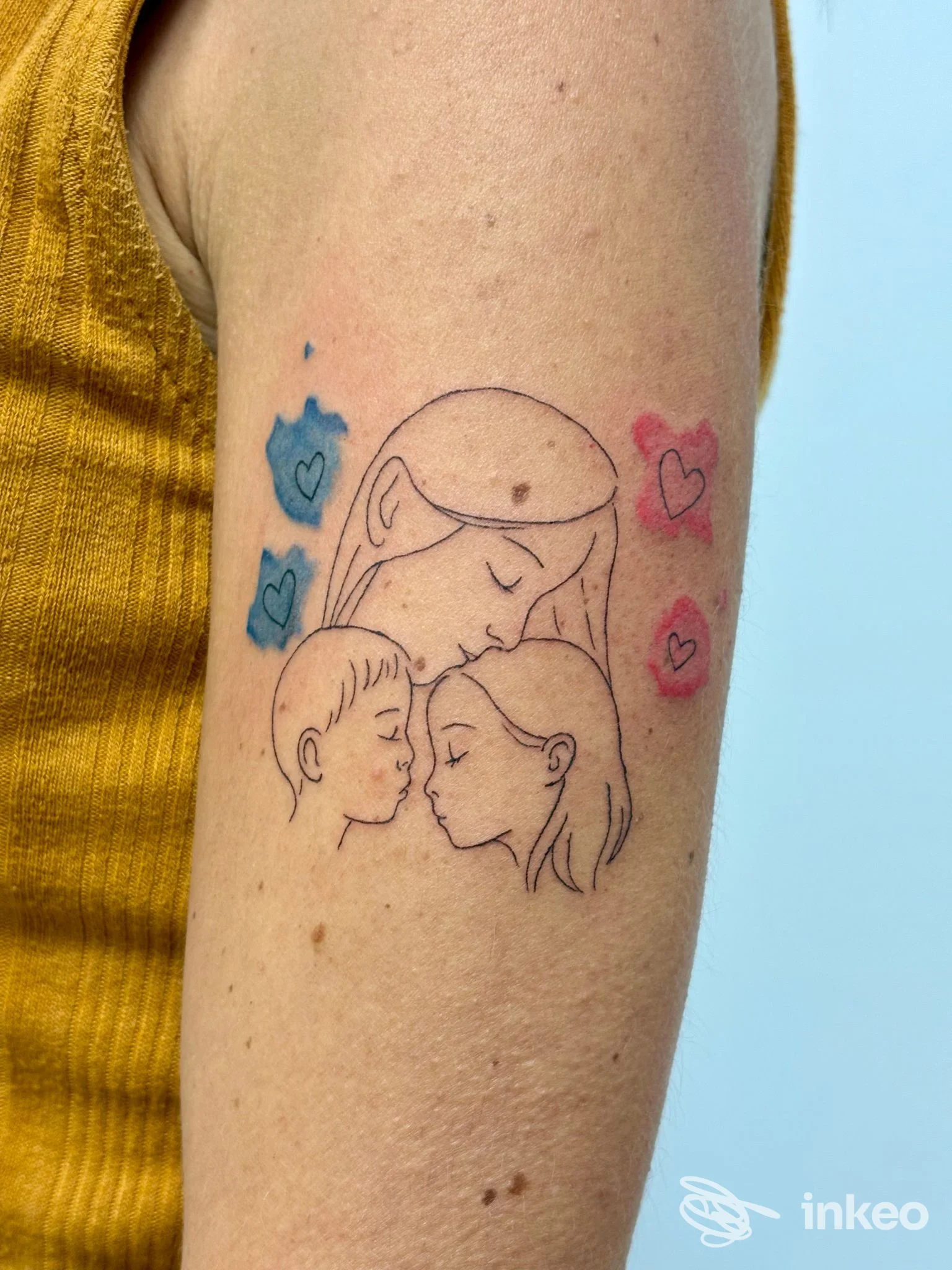Tatuaggio lineare di madre e due bambini sul braccio superiore, con dettagli colorati blu e rosa. EleTattoo, Lanusei.