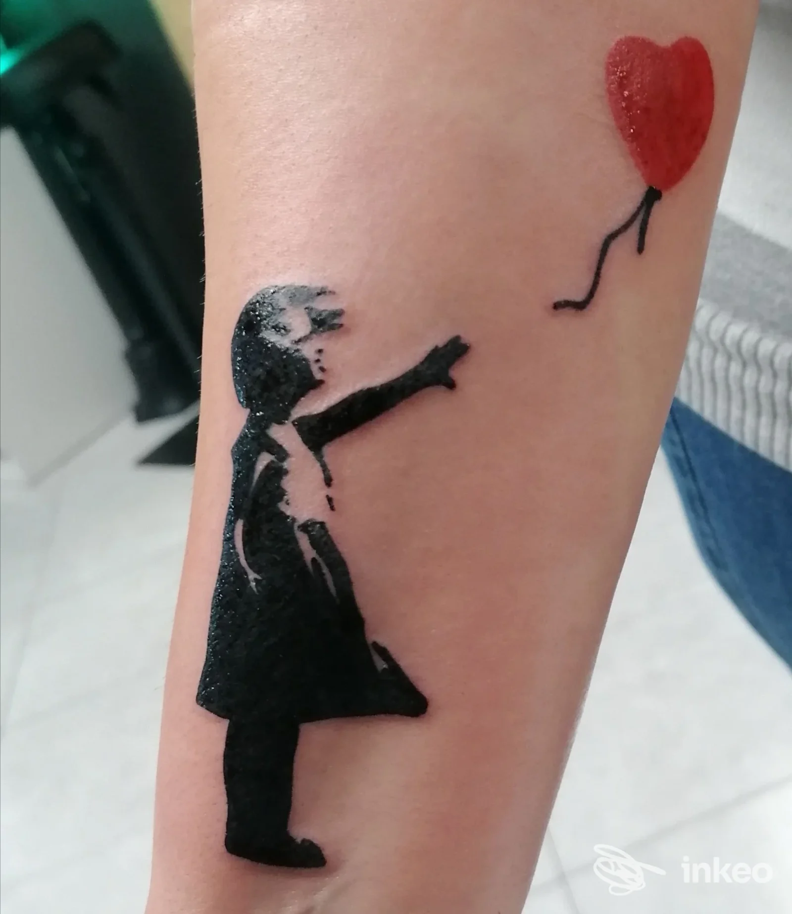 Tatuaggio su avambraccio con bambina stilizzata nera che indica un palloncino a cuore rosso — Eternity 17 Tattoo Shop, Selargius.