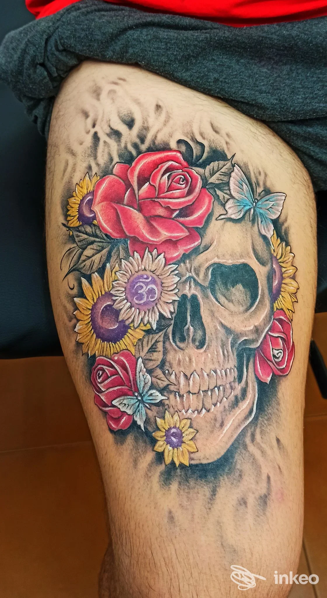 Tatuaggio a colori sulla coscia con teschio centrale circondato da rose rosse, girasoli gialli e farfalle azzurre. GD Tattoo, Terralba.