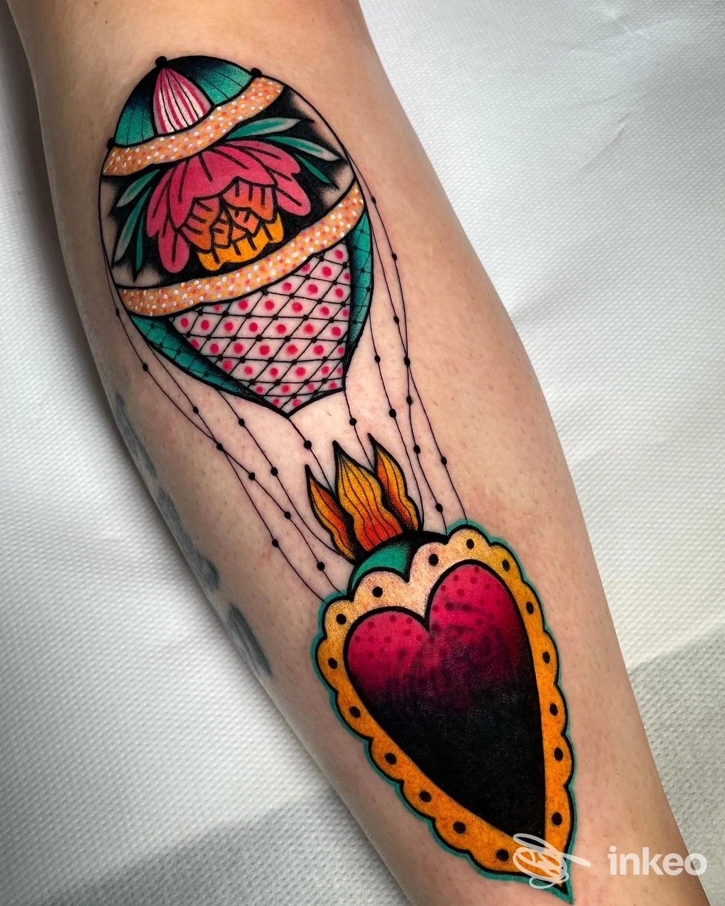 Tatuaggio colorato sull'avambraccio sinistro: cuore nero con contorno giallo e un grande palloncino decorato collegato tramite corde, su sfondo chiaro.