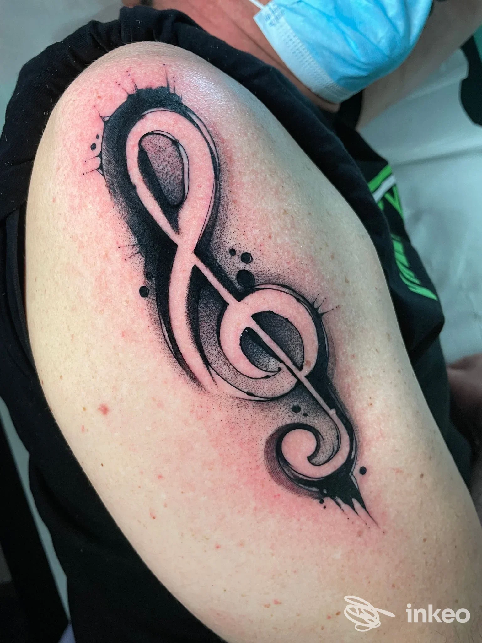 Tattoo visibile sulla pelle del braccio: una chiave di violino con sfumature nere e sfondo rosa, realizzata in stile realistico/watercolor.
