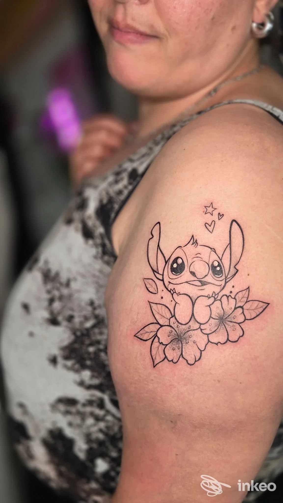 Tatuaggio linework di Stitch su fiori sul braccio superiore; foto fornita da Kind of Magic Tattoo Studio, Cagliari.