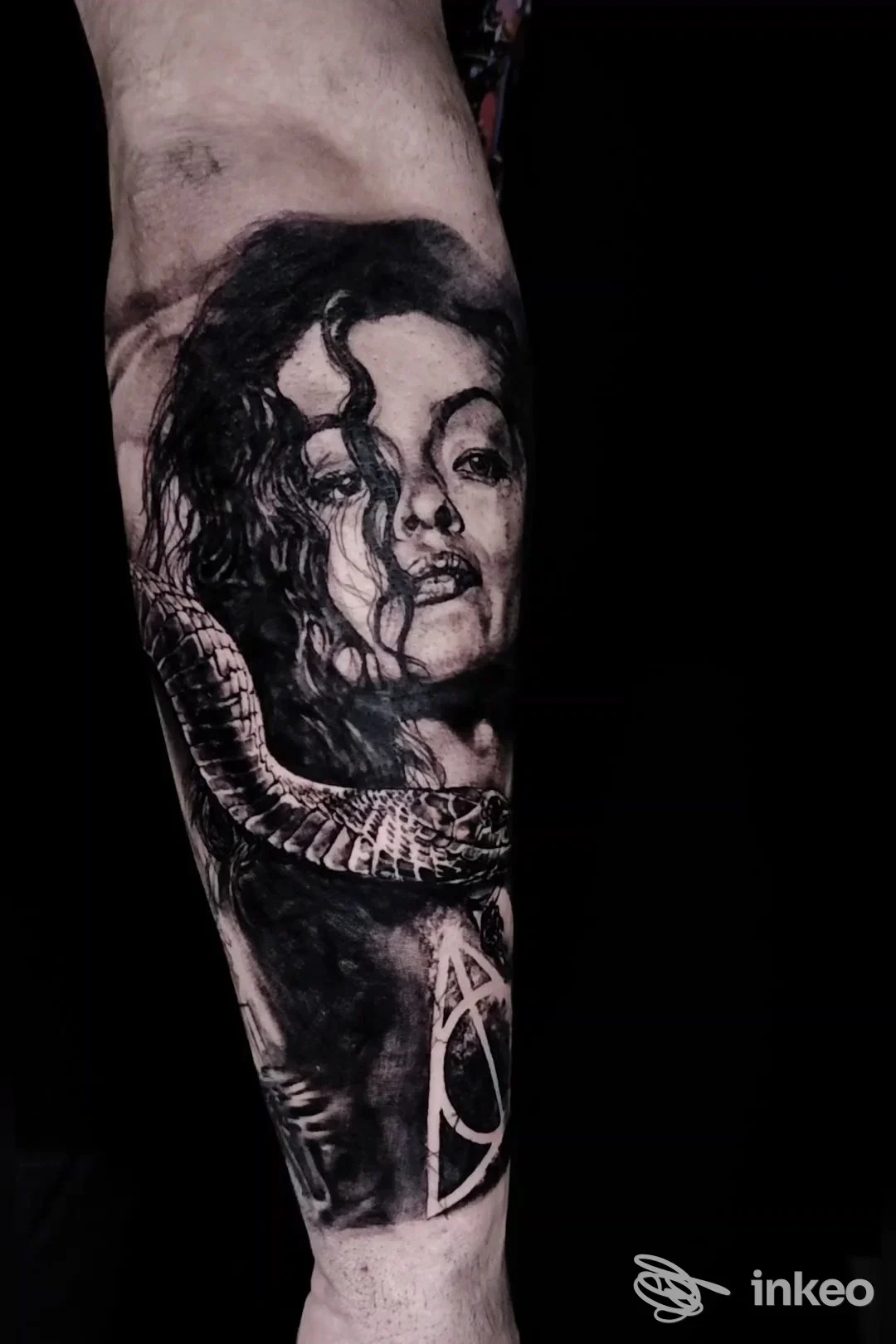 Tattoo in bianco e nero su avambraccio raffigurante un volto femminile avvolto da una serpente; corpo visibile è pelle reale, contesto minimo.