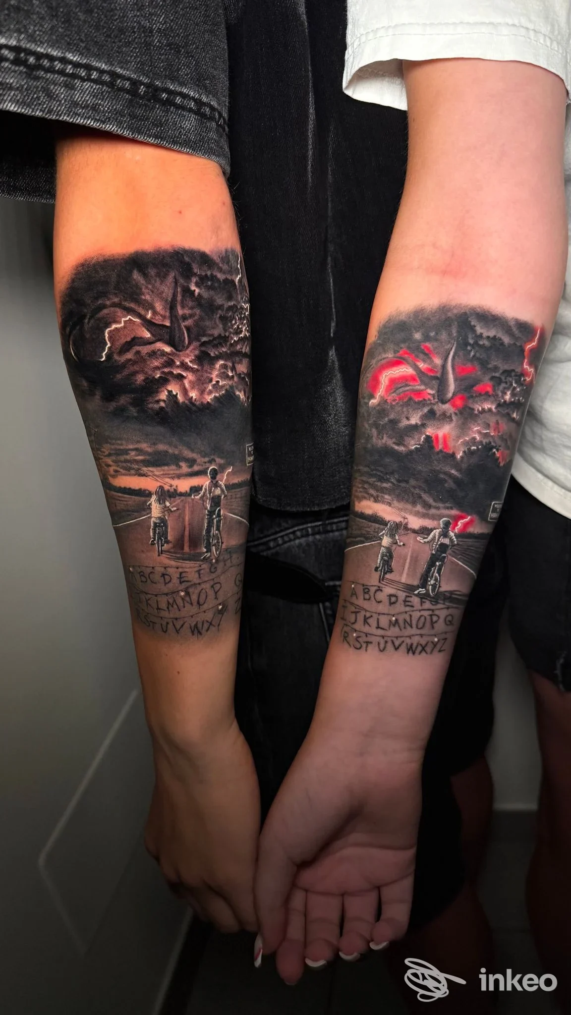 Due avambracci vicini mostrano tatuaggi realistici in tonalità nere e grigie con dettagli di paesaggio e figure umane; sull’avambraccio destro si notano accenti rossi.