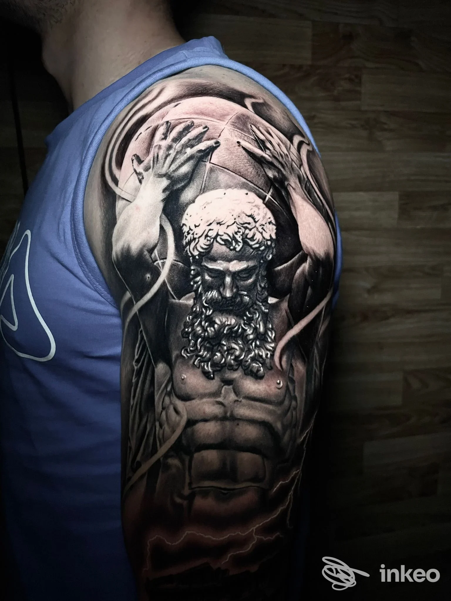Tattoo realistico sullupper arm sinistro: figura barbuta muscolosa con braccia sollevate; pelle visibile, nessun volto umano intero, scatto singolo.