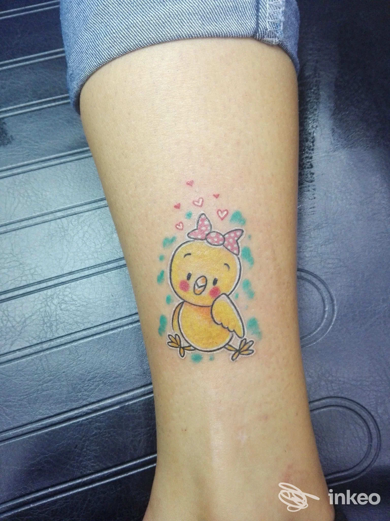 Tatuaggio sul polpaccio di un pulcino cartoon con fiocco e cuoricini, colori giallo, rosa e turchese — Black Stones Ink, Santa Teresa di Riva.