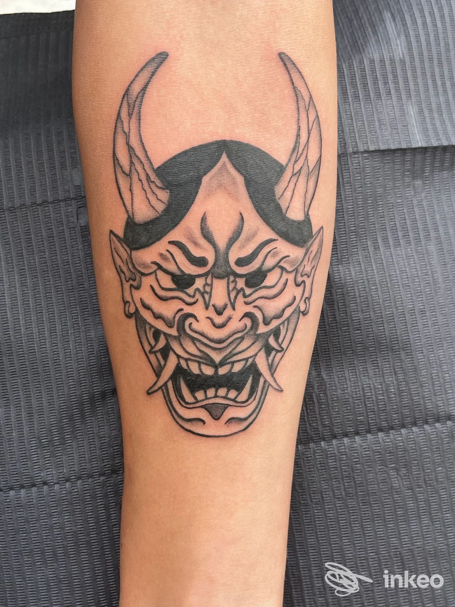 Tatuaggio a inchiostro nero di una maschera demoniaca sull'avambraccio, foto fornita da Basilisk Tattoo Studio, Terrasini.