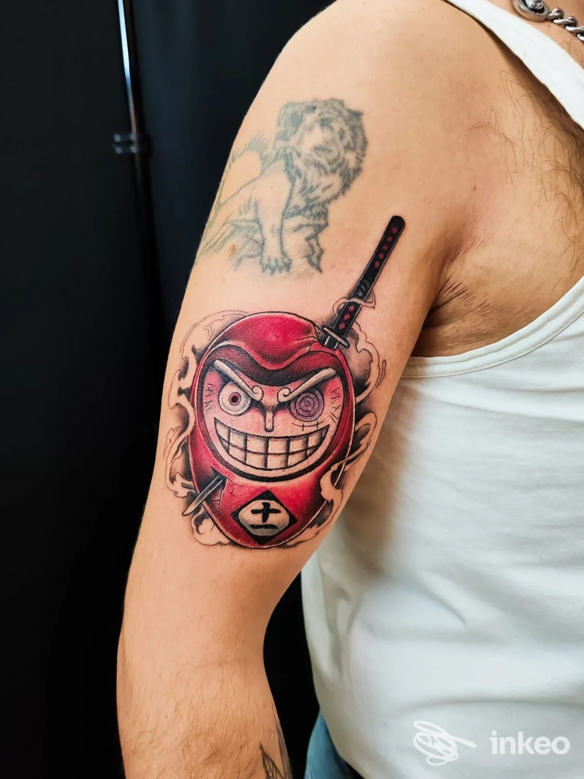 Tatuaggio colorato a forma di maschera rossa con katana sul braccio superiore; foto fornita da Fuoriposto Tattoo, Trapani.