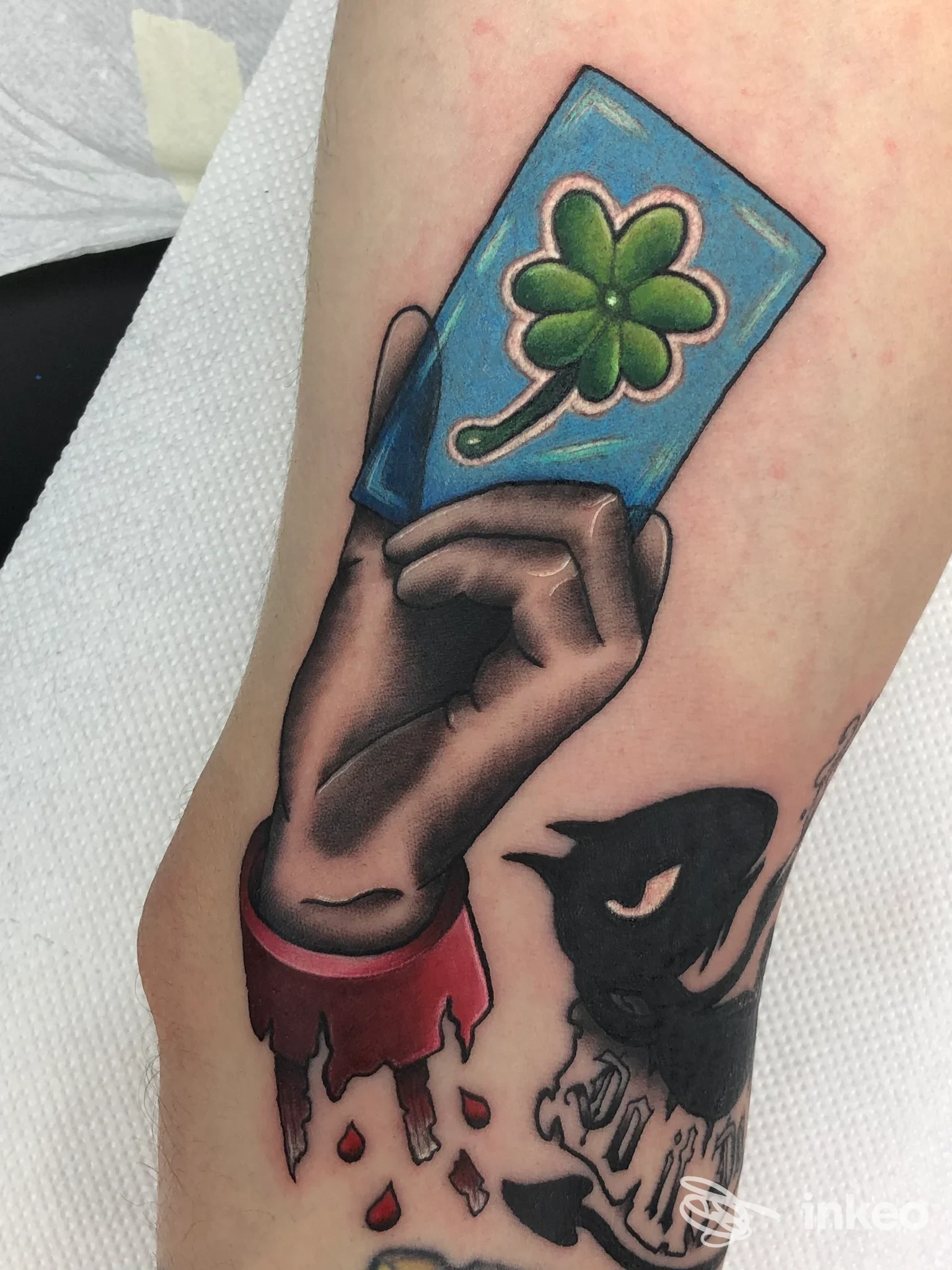 Tatuaggio su avambraccio: mano stilizzata che tiene una carta con quadrifoglio, con corvo nero e effetto sangue. Ghost Lost tattoo shop, Bagheria.