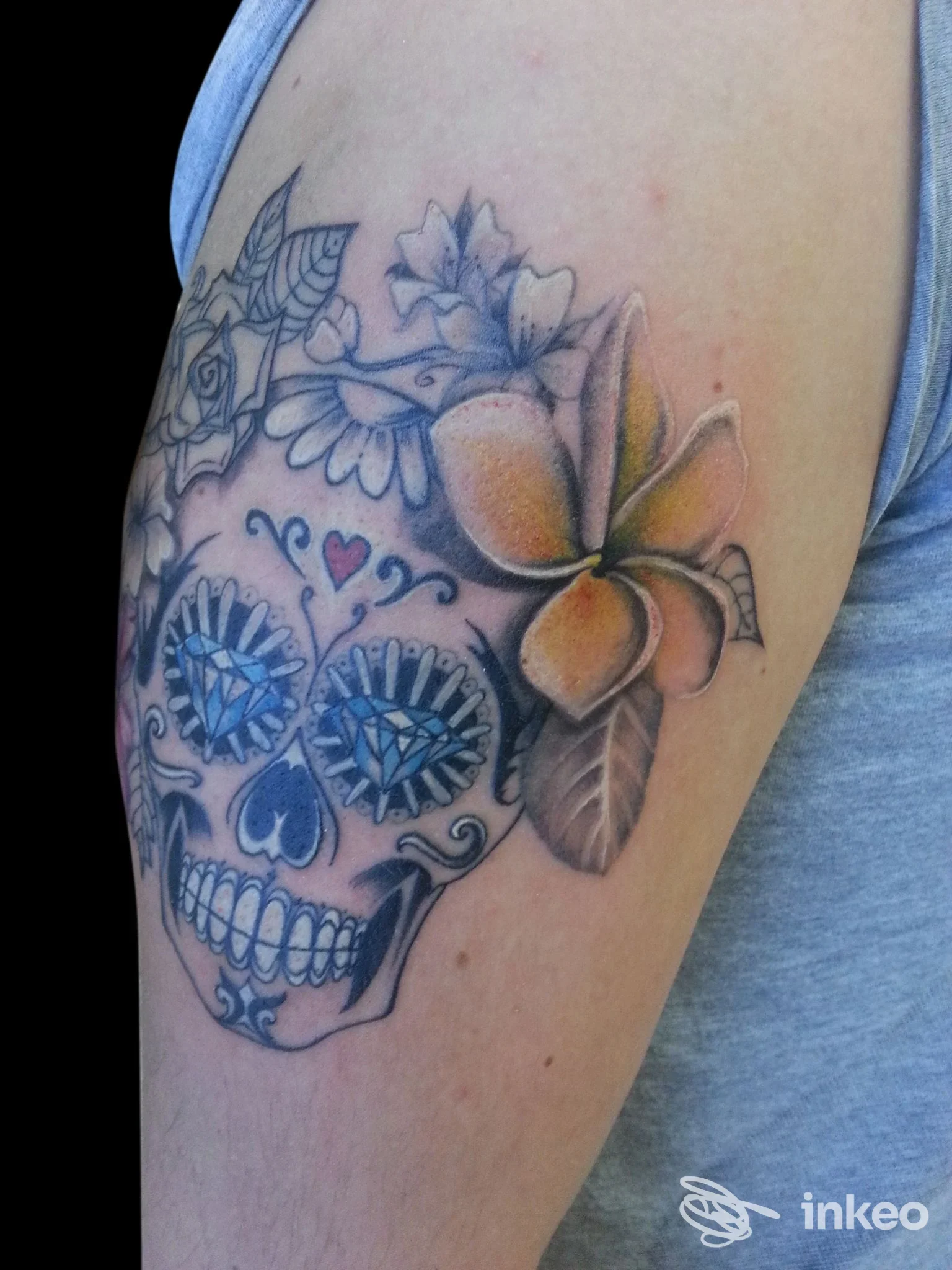 Tatuaggio visibile sulla pelle del braccio: una sugar skull decorata con fiori, colori blu e arancione, scultura dettagliata su pelle nuda.