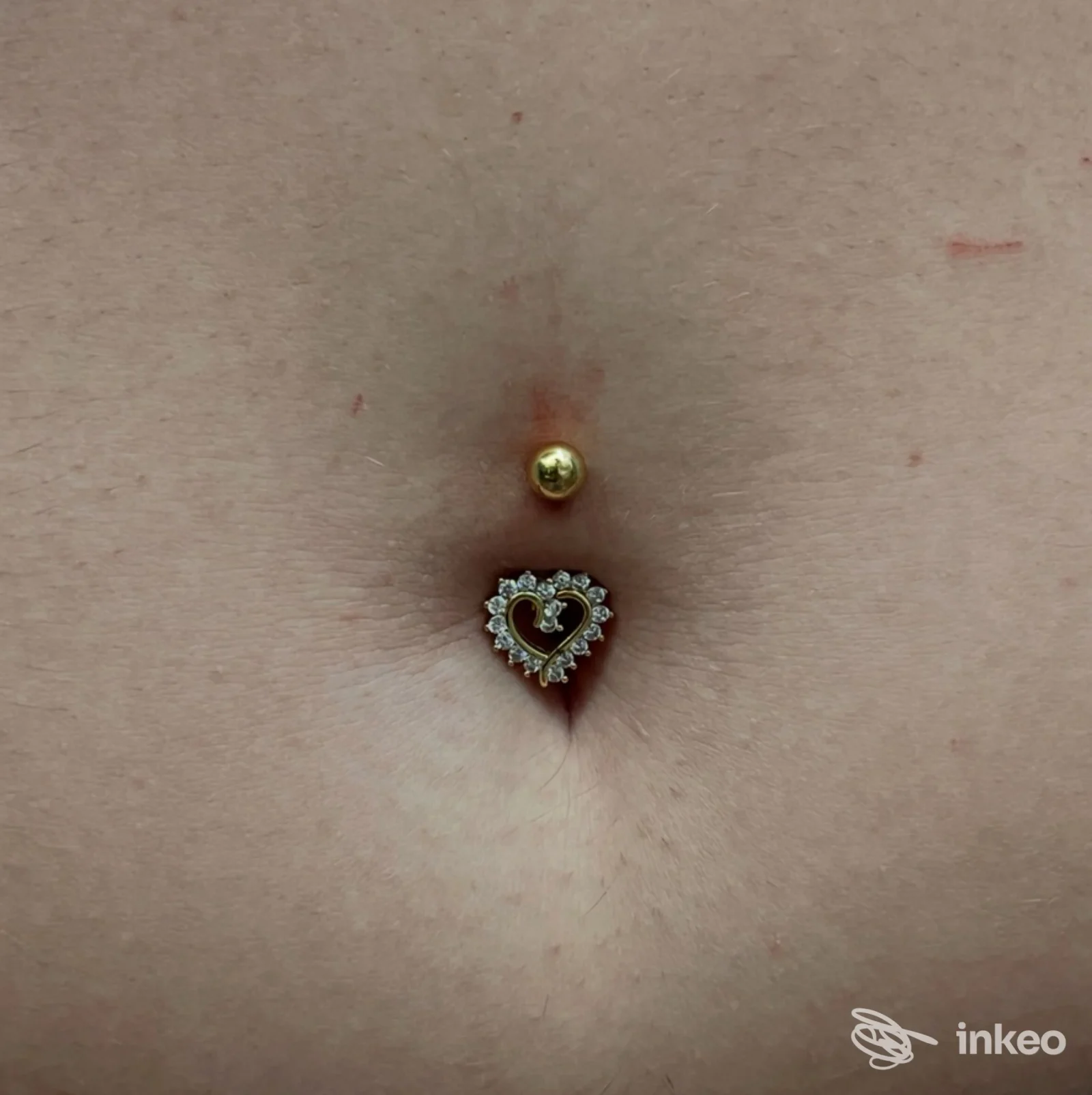 Primo piano di un piercing ombelicale con sfera dorata e ciondolo a cuore con zirconi, foto fornita da Piercing house presso Vanity Mara a Sciacca.
