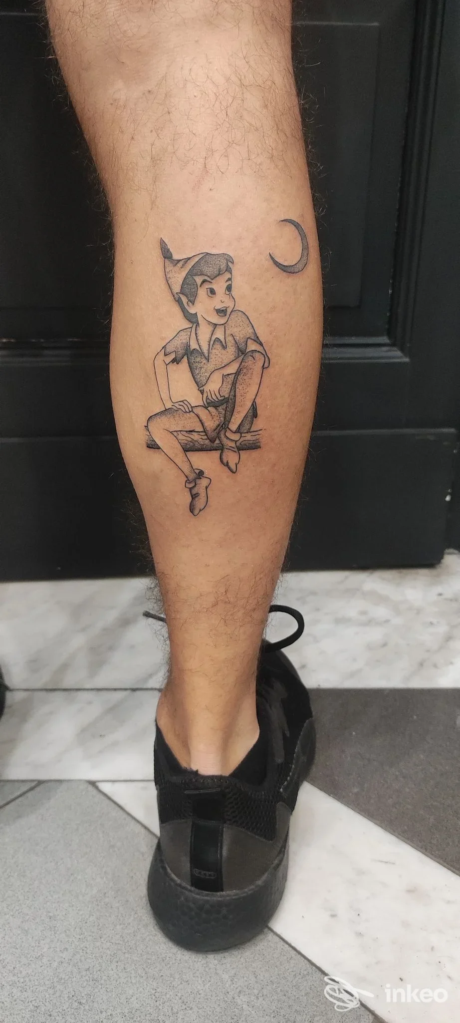 Tatuaggio in bianco e nero di un personaggio seduto e una mezza luna sul polpaccio, Sideshow Studio Piercing & Tattoo, Catania.