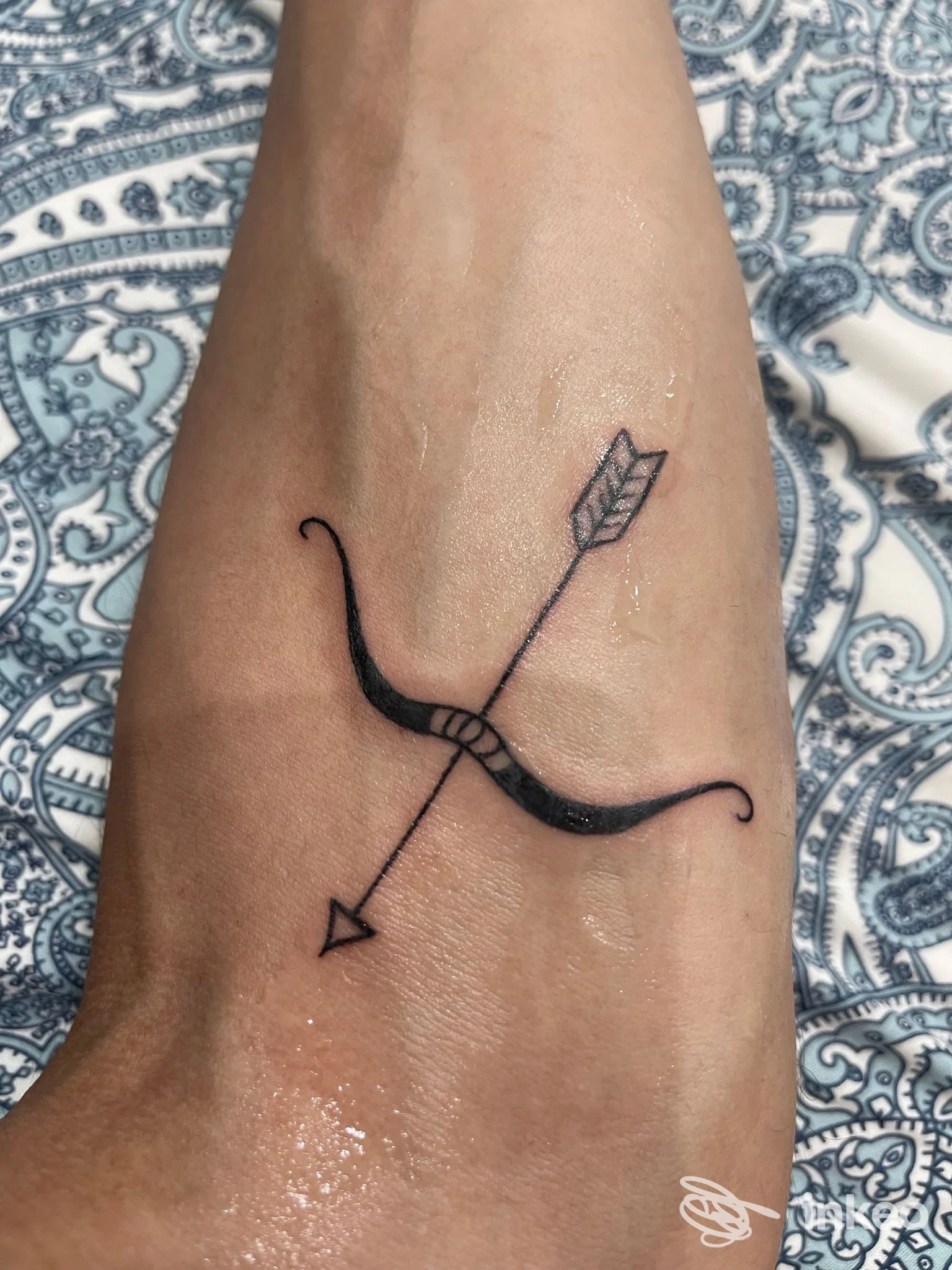 Tatuaggio minimal nero di arco e freccia sul dorso del piede, inviato da Skin Tattoo di La Rocca Adriano, Giardini-Naxos.