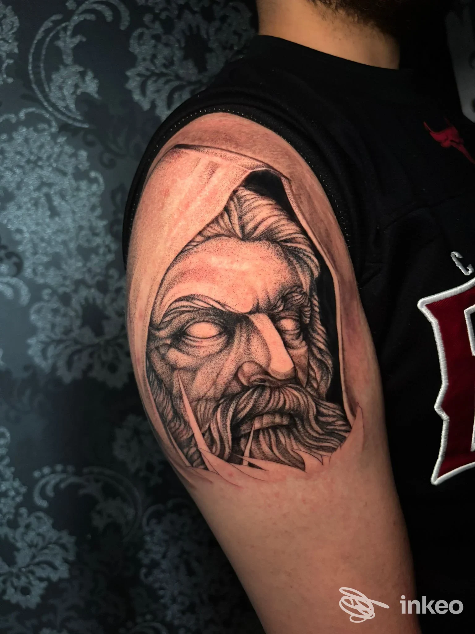 Tatuaggio realistico in bianco e nero di un volto con cappuccio sul deltoide, fotografia per Studio Tattoo & Piercing Iracema Gravina di Catania.