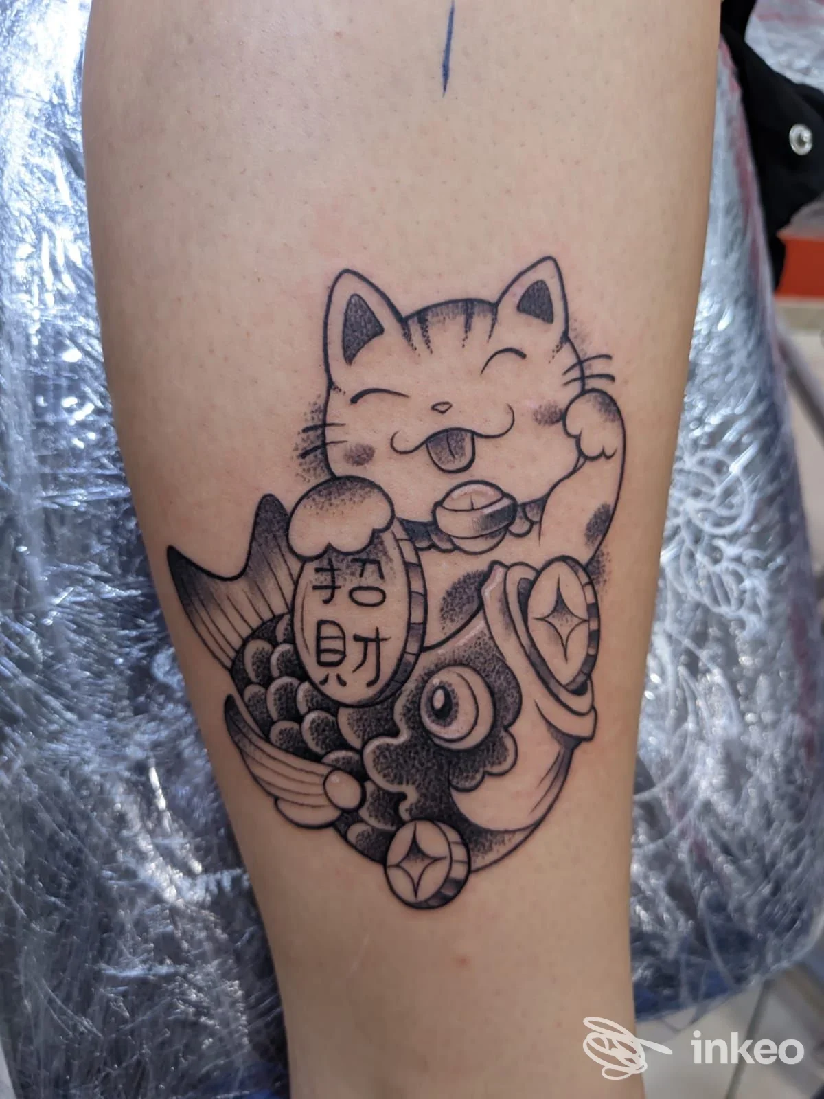 Tatuaggio in nero e grigio di un maneki-neko su un pesce sul polpaccio, Tattoo Studio di Stefano Pennacchio, Lentini.