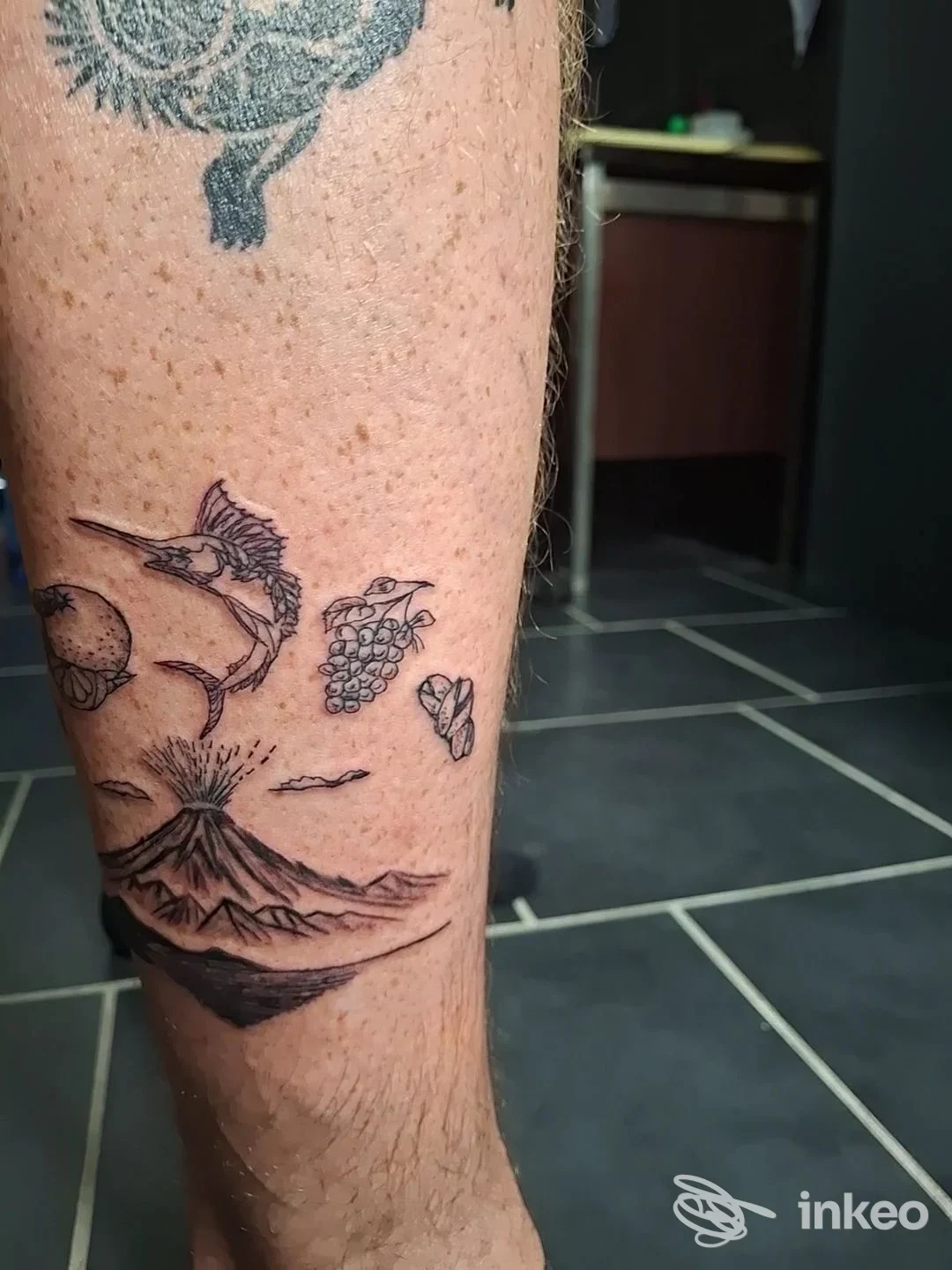 Dettaglio di tatuaggi in bianco e nero sul polpaccio, tra cui un vulcano eruttante e un marlin; Vali Tattoo & Bodypiercing, Catania.