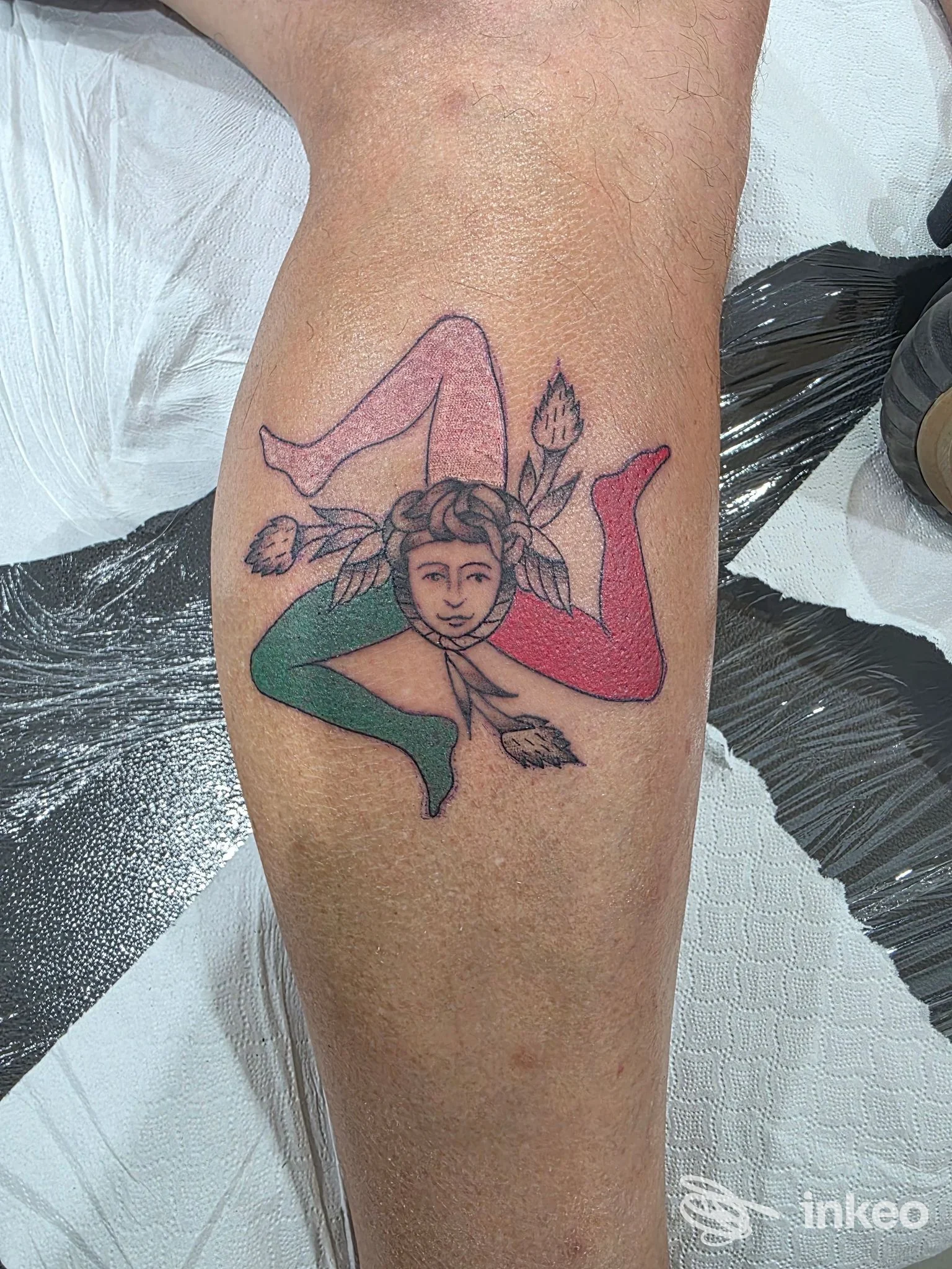 Tatuaggio sul polpaccio raffigurante una figura stilizzata con volto centrale e gambe colorate; Vintage tattoo sopra il porto, Terrasini.