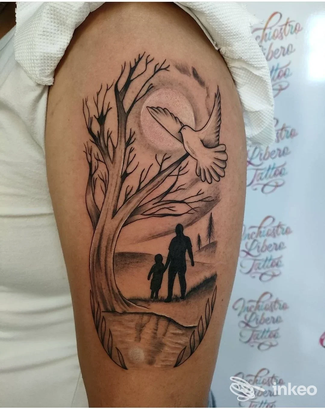 Tatuaggio black & grey su braccio superiore con albero, colomba in volo e due figure in silhouette. Immagine fornita da Inchiostro Libero Studio Tattoo, Partinico.