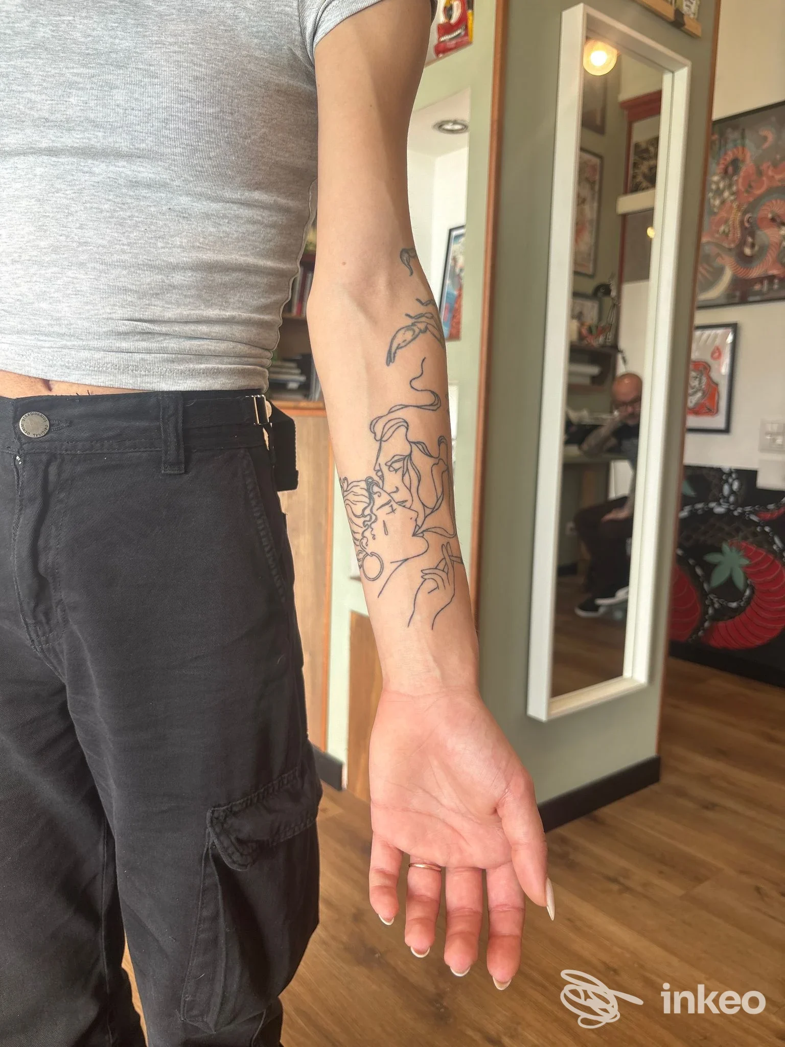 Tattoo sull’avambraccio sinistro con linee nere; l’immagine è scattata in un salone di tatuaggi, sfondo con decorazioni e mobili, nessun volto visibile.