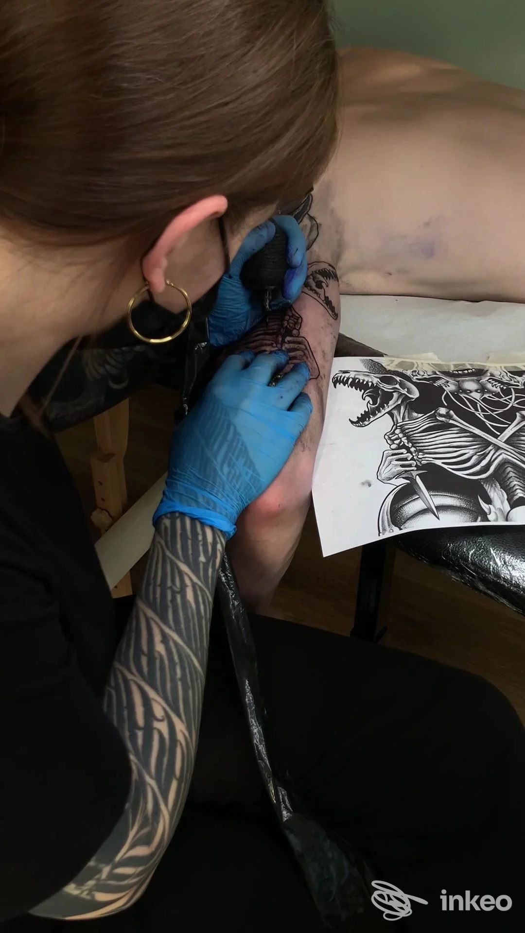 Un'operatrice tatuatrice con guanti blu lavora sull’avambraccio di una cliente, mostrando strumenti e un disegno in bianco e nero su carta accanto alla pelle.