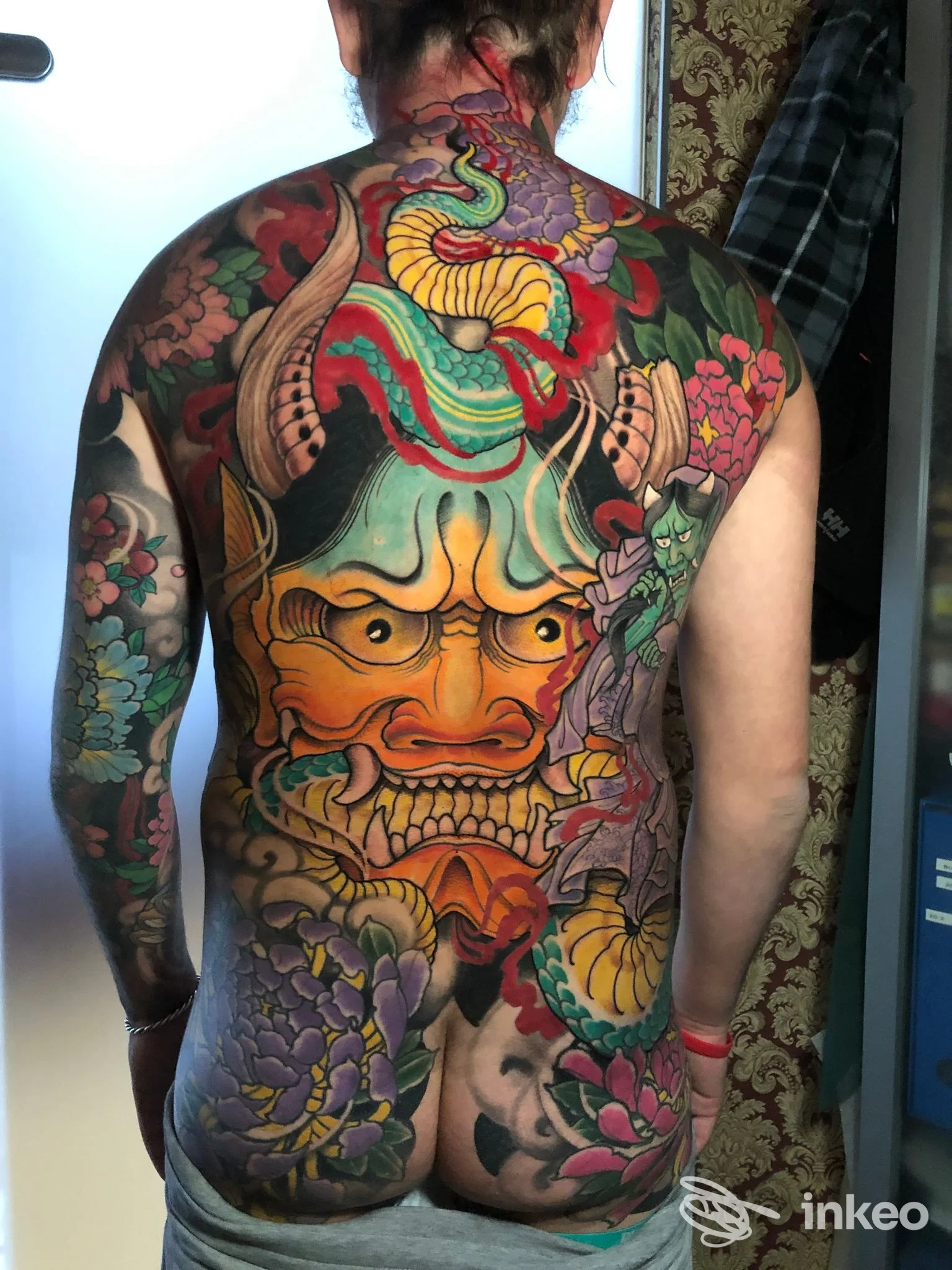Ampia schiena tatuata con un grande volto demoniaco e motivi floreali in colori vividi, foto fornita da Gentes Scriptae Tattoo & Piercing Studio, Firenze.