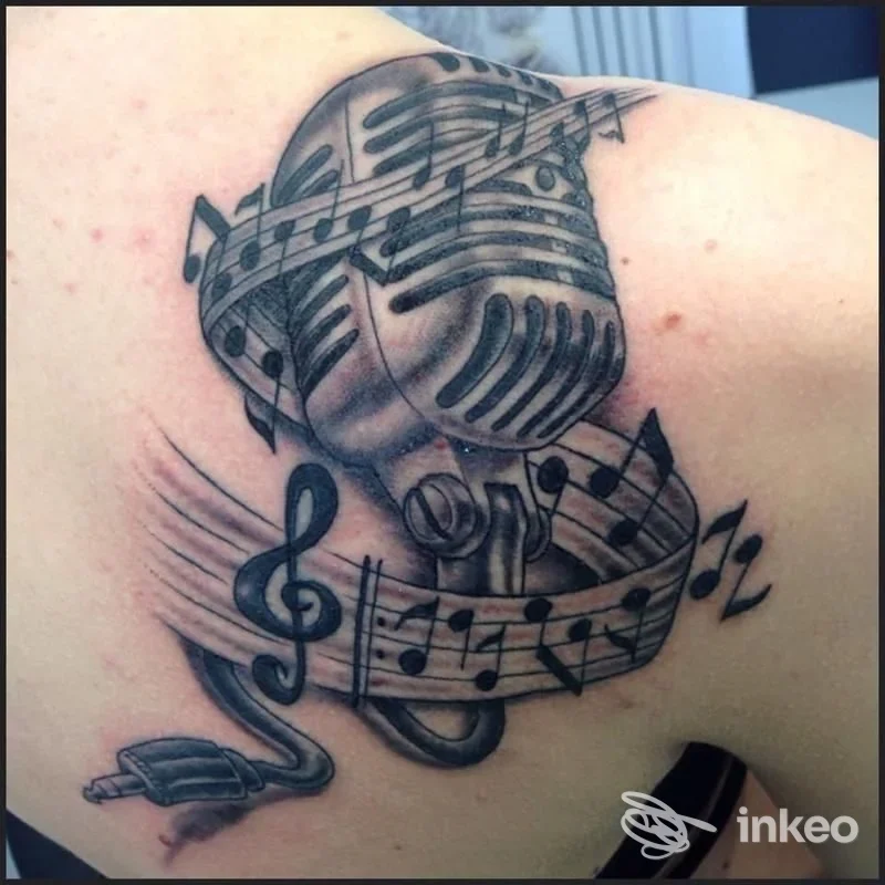 Tatuaggio microfono vintage avvolto da spartito musicale sul deltoide, realizzato da Gianni Tattoo Studio a Rosignano Solvay-Castiglioncello.