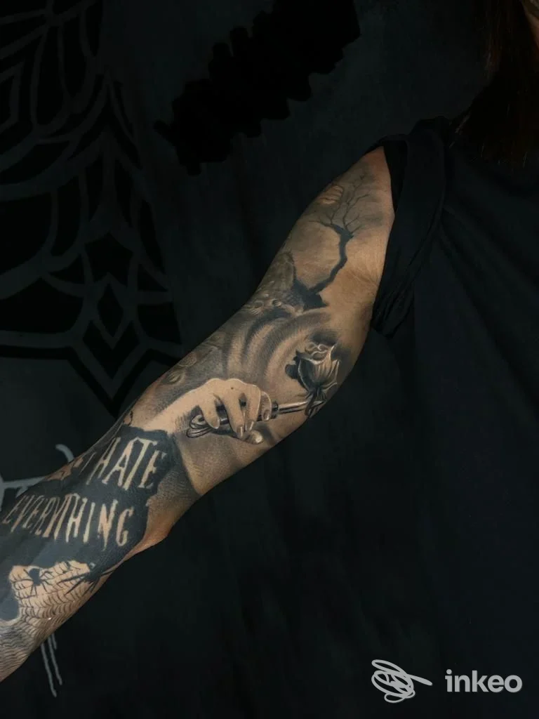 Avambraccio visibile con tatuaggio complesso, raffigurante una mano e altri motivi su sfondo scuro; nessun volto umano visibile; immagine singola presa in studio.