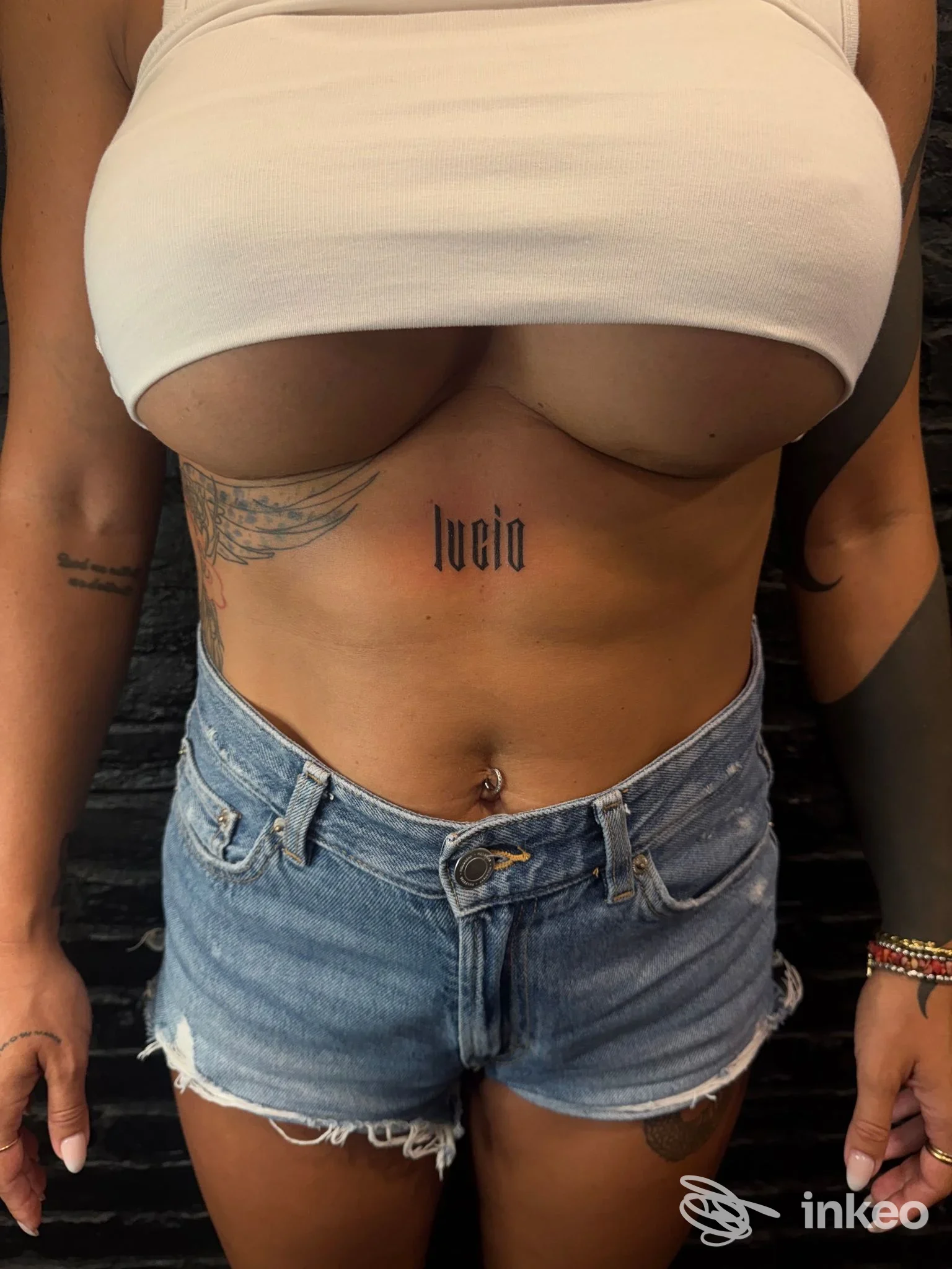 Primo piano del torso con tatuaggio script sullo sterno, ali laterali e piercing all'ombelico, fotografato per GIULS TATTOOER a Follonica.